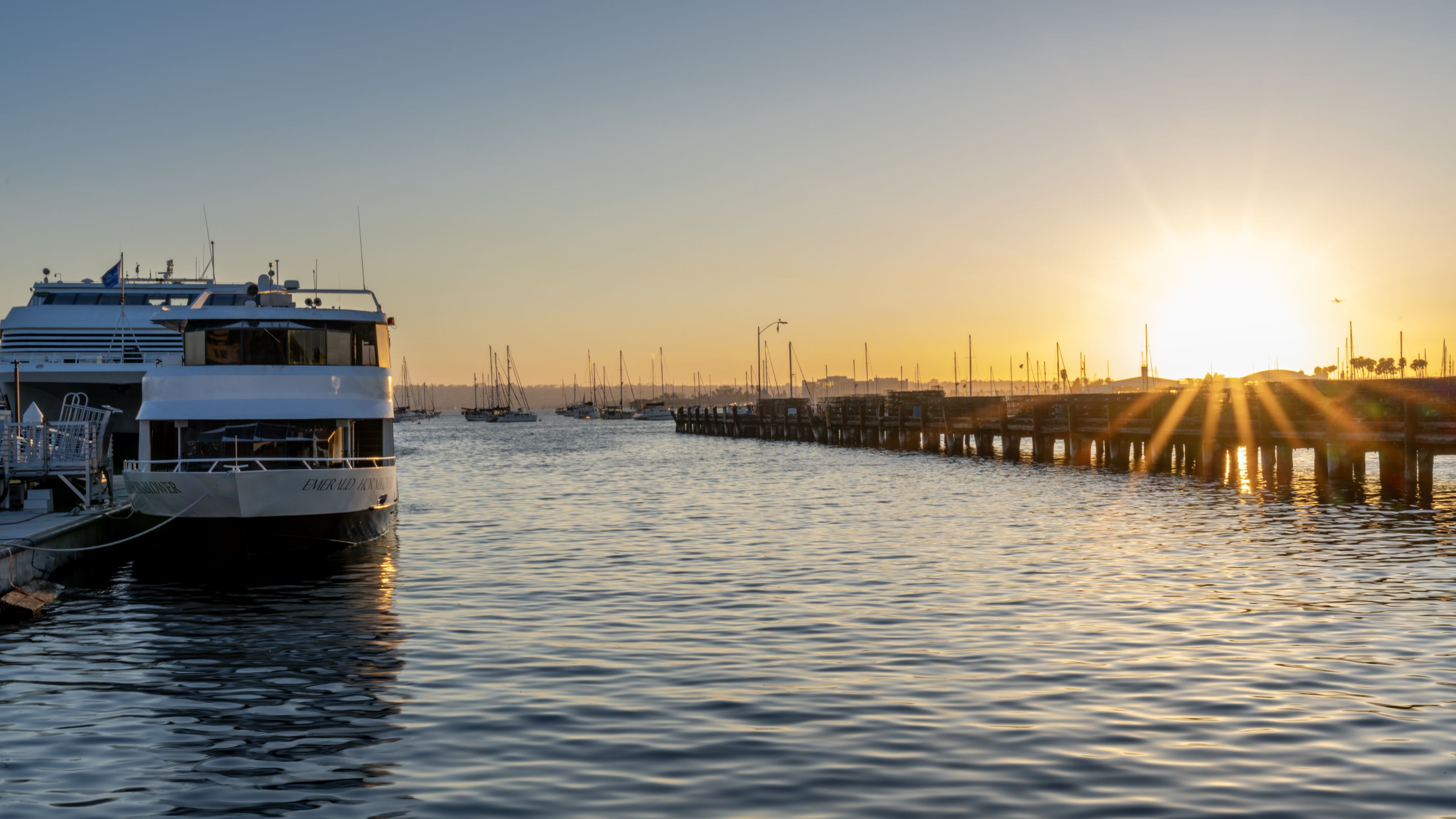 A boat is docked at a pier with the sun setting over a calm harbor and distant sailboats.