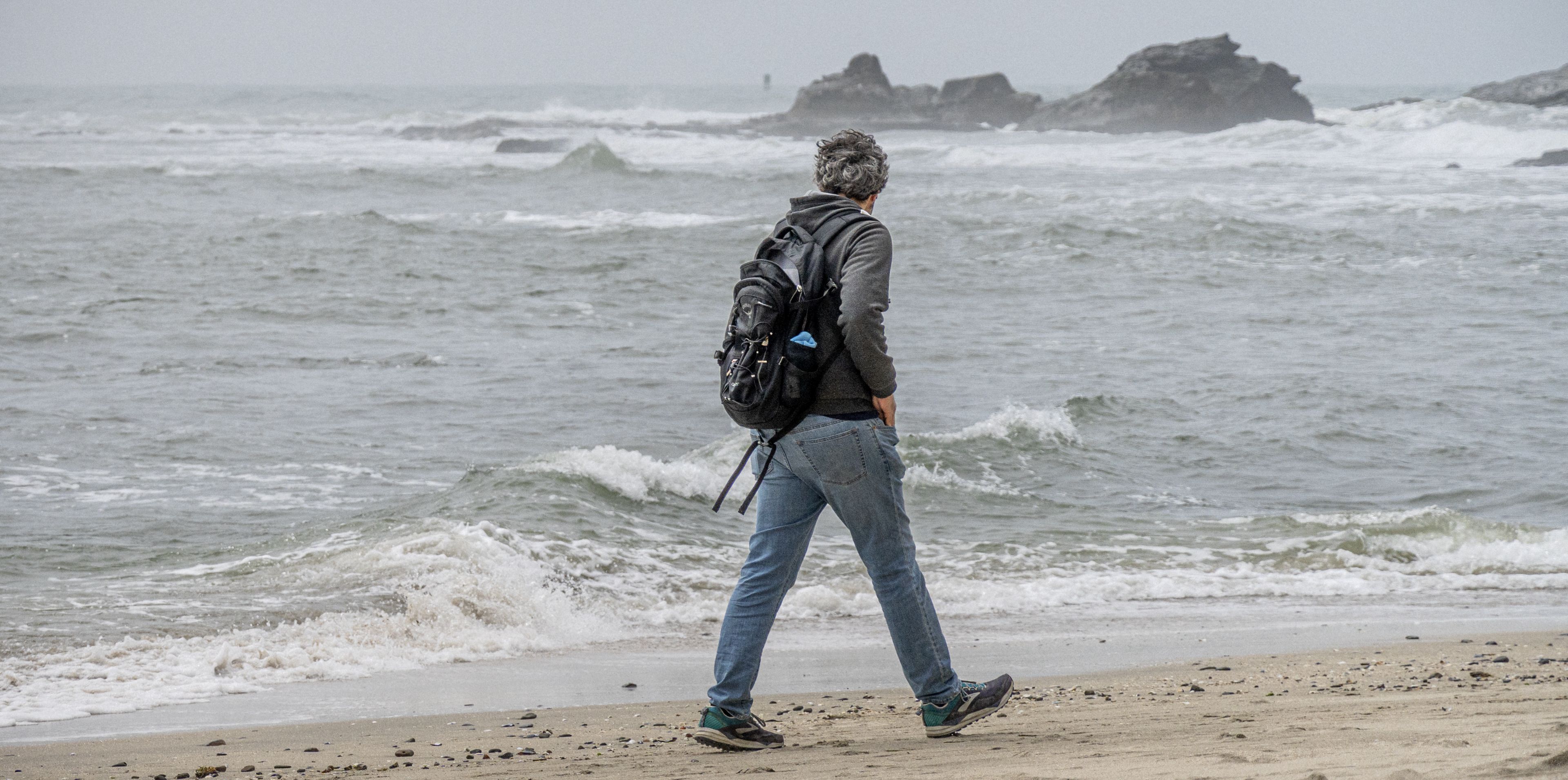 An individual with a backpack walks along the shore with waves in the background.