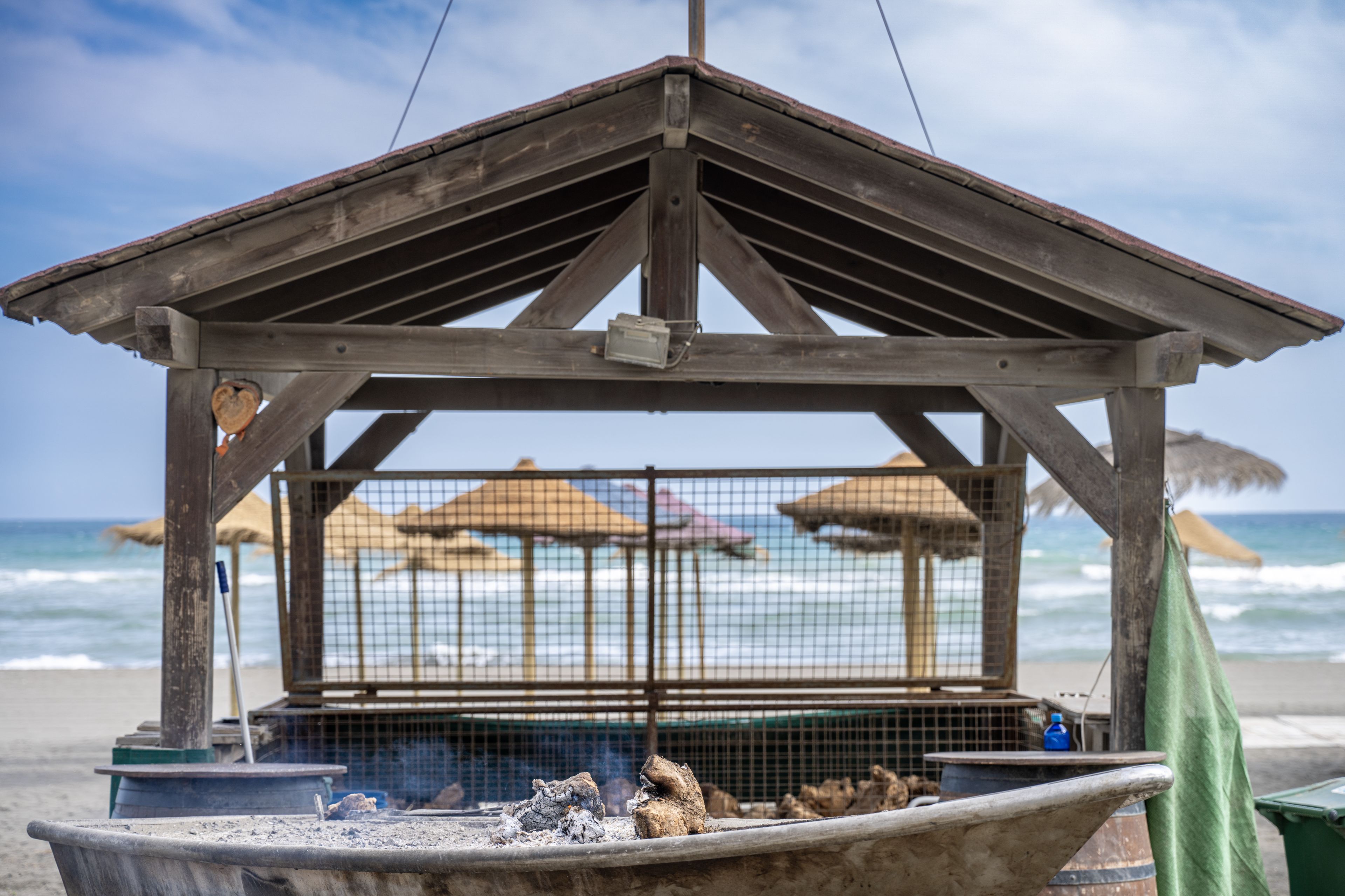 A wooden pavilion near the beach with straw umbrellas and a grill in the foreground.