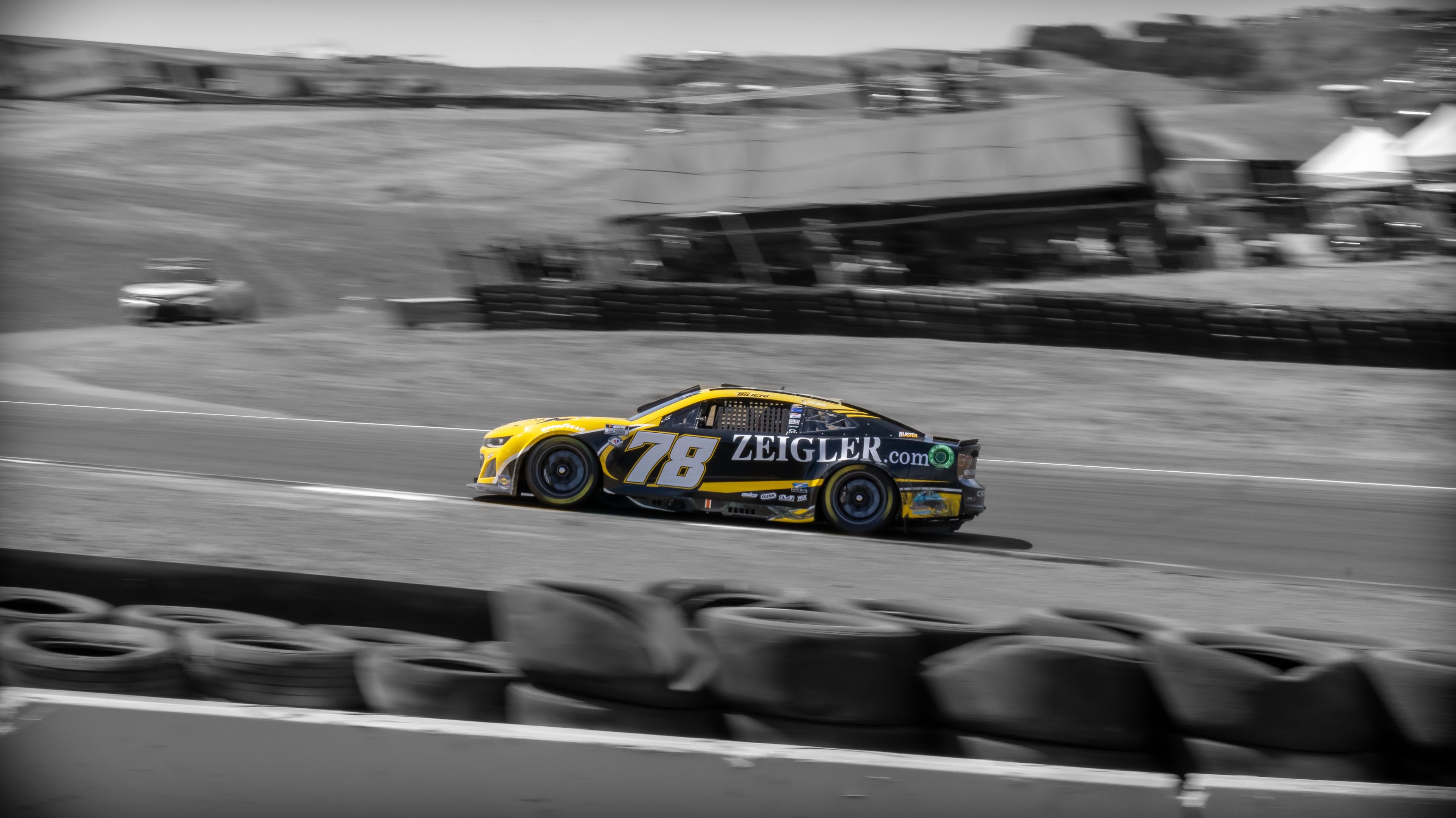 A yellow racing car speeds around a track with tire barriers in the foreground.