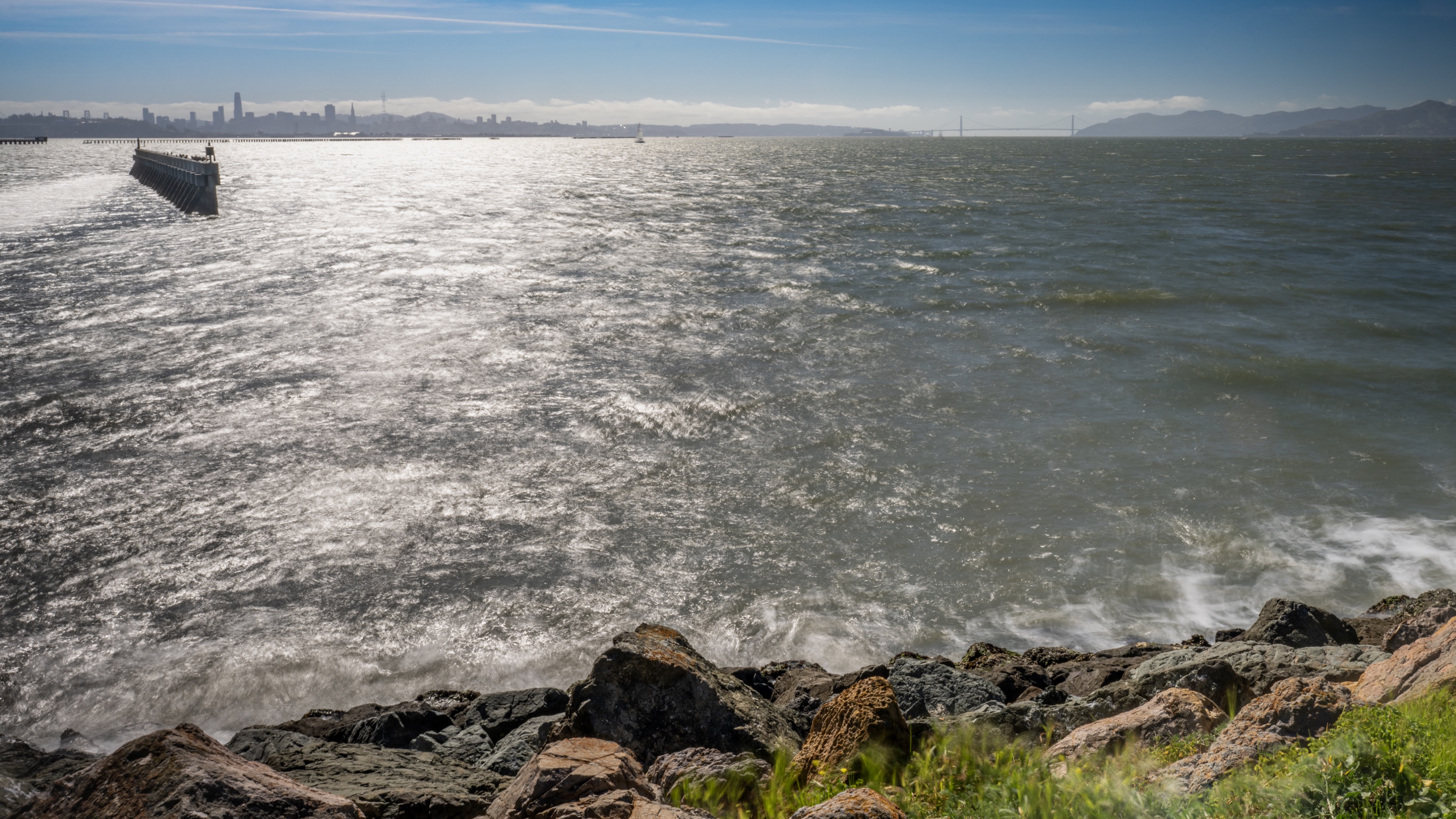 A rocky shoreline with waves lapping against the stones and a distant city skyline.