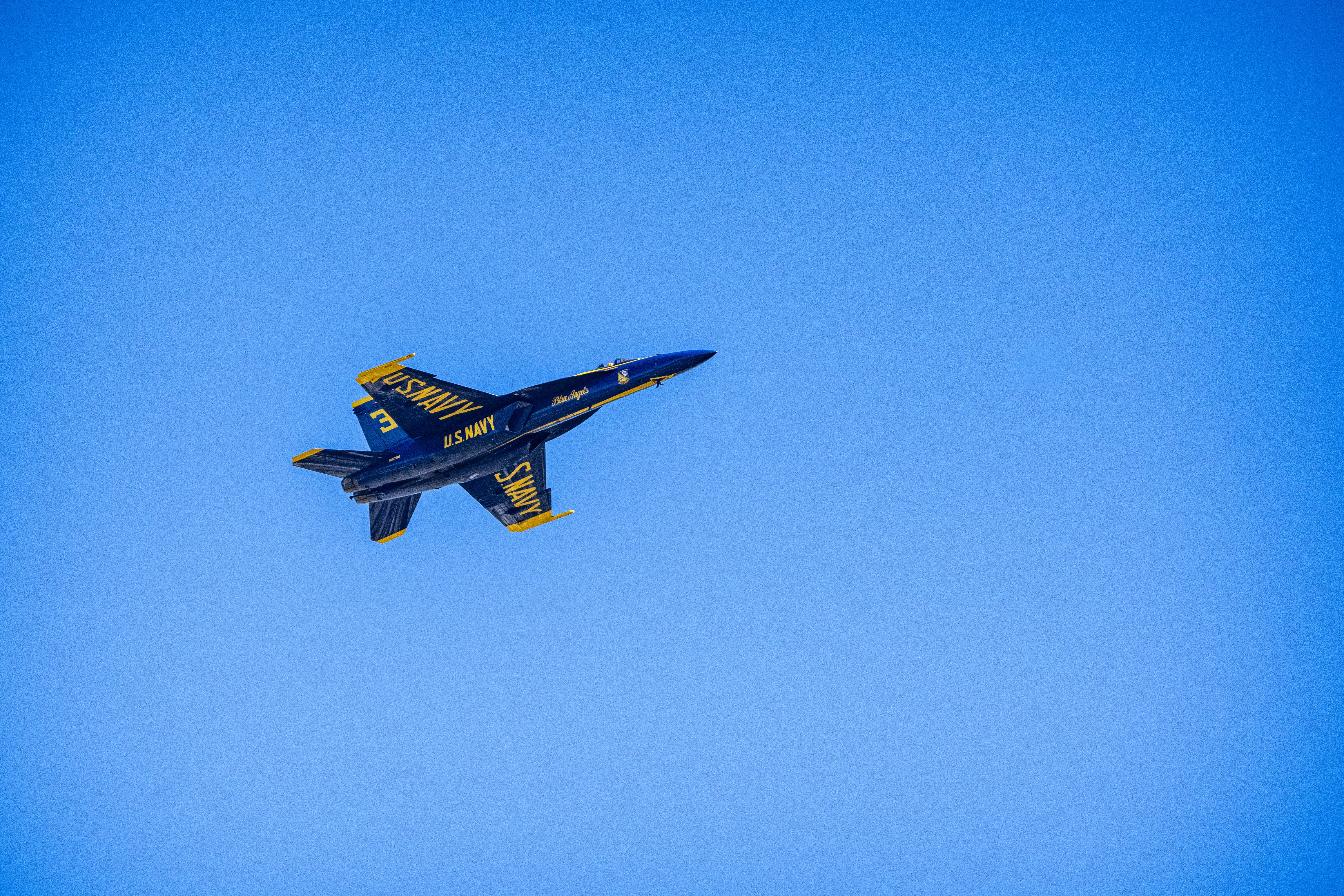 A Blue Angels jet banks across the sky with a faint smoke trail behind it.