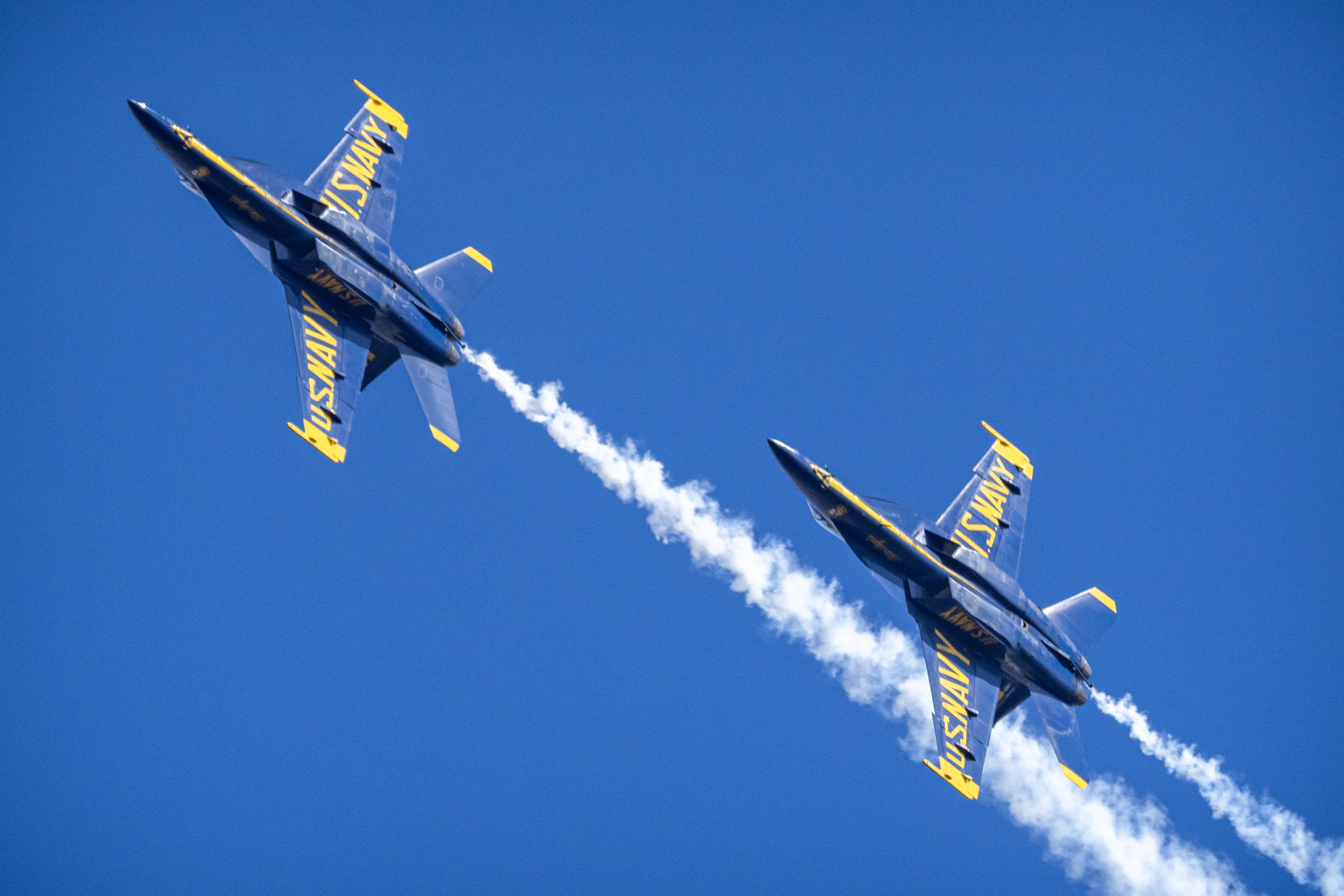 Two Blue Angels jets fly close together with smoke trailing behind them.