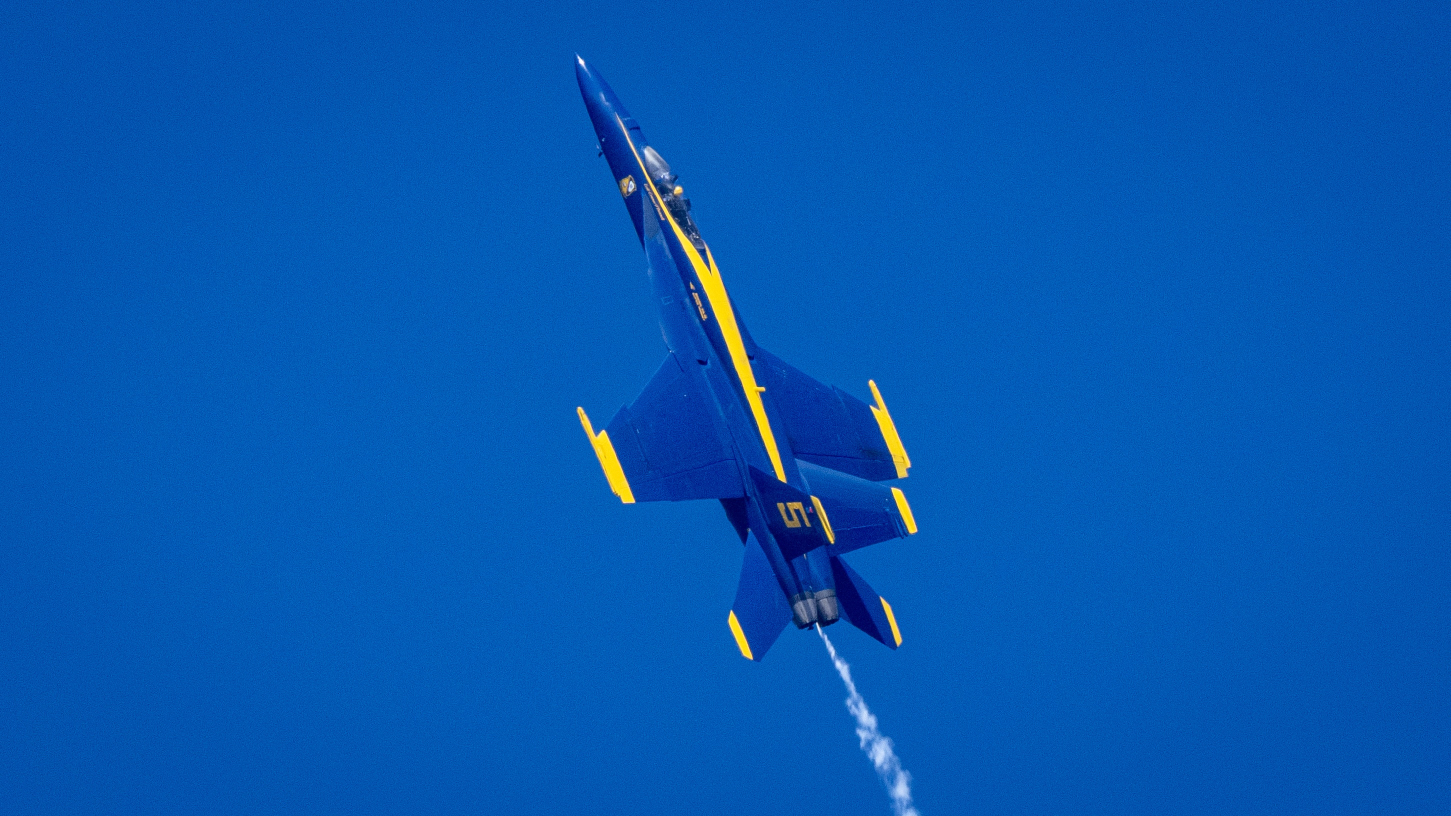 The underside of a Blue Angels jet passes against a clear sky.