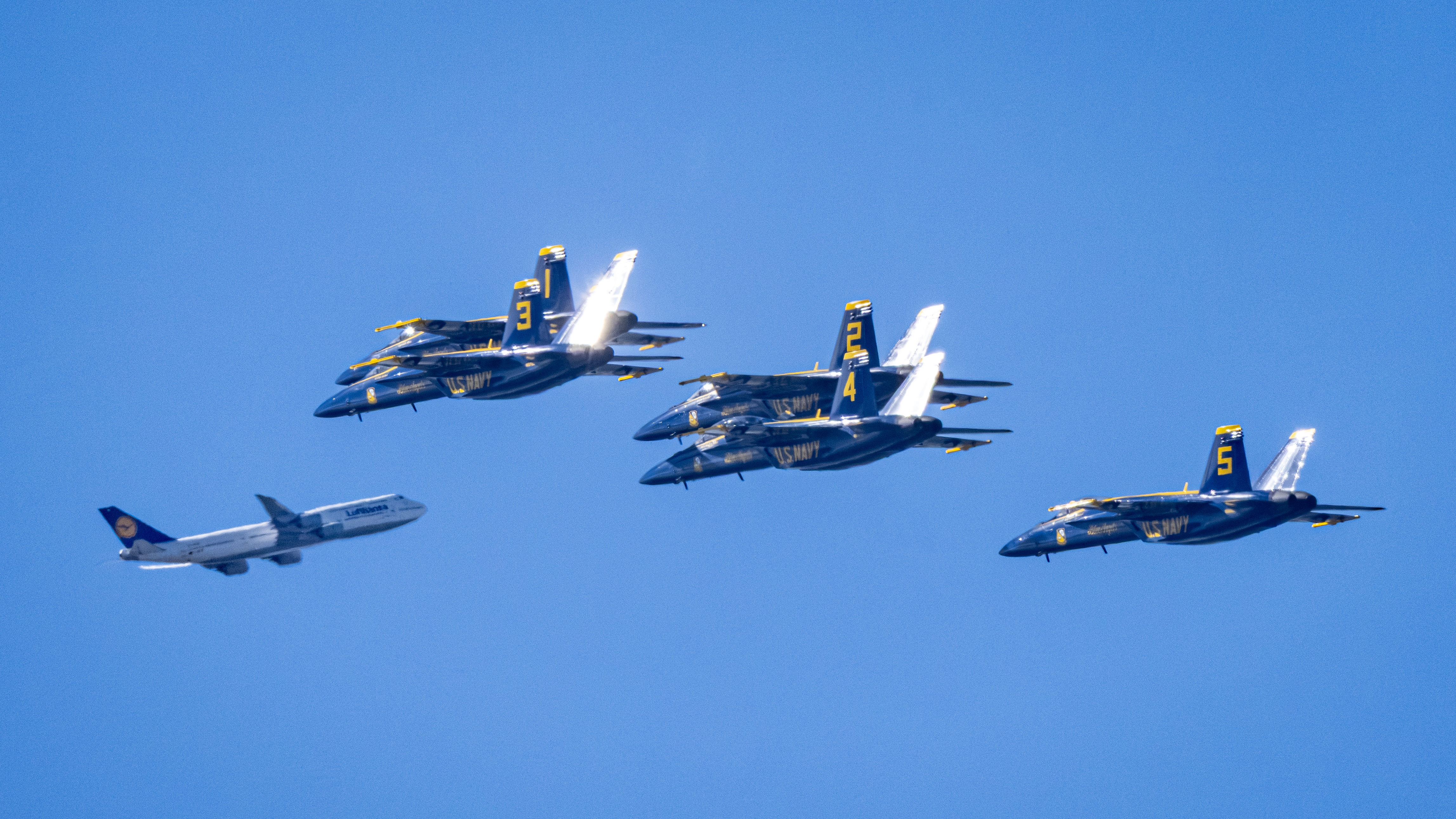 Five Blue Angels jets fly in formation beside a gray aircraft.