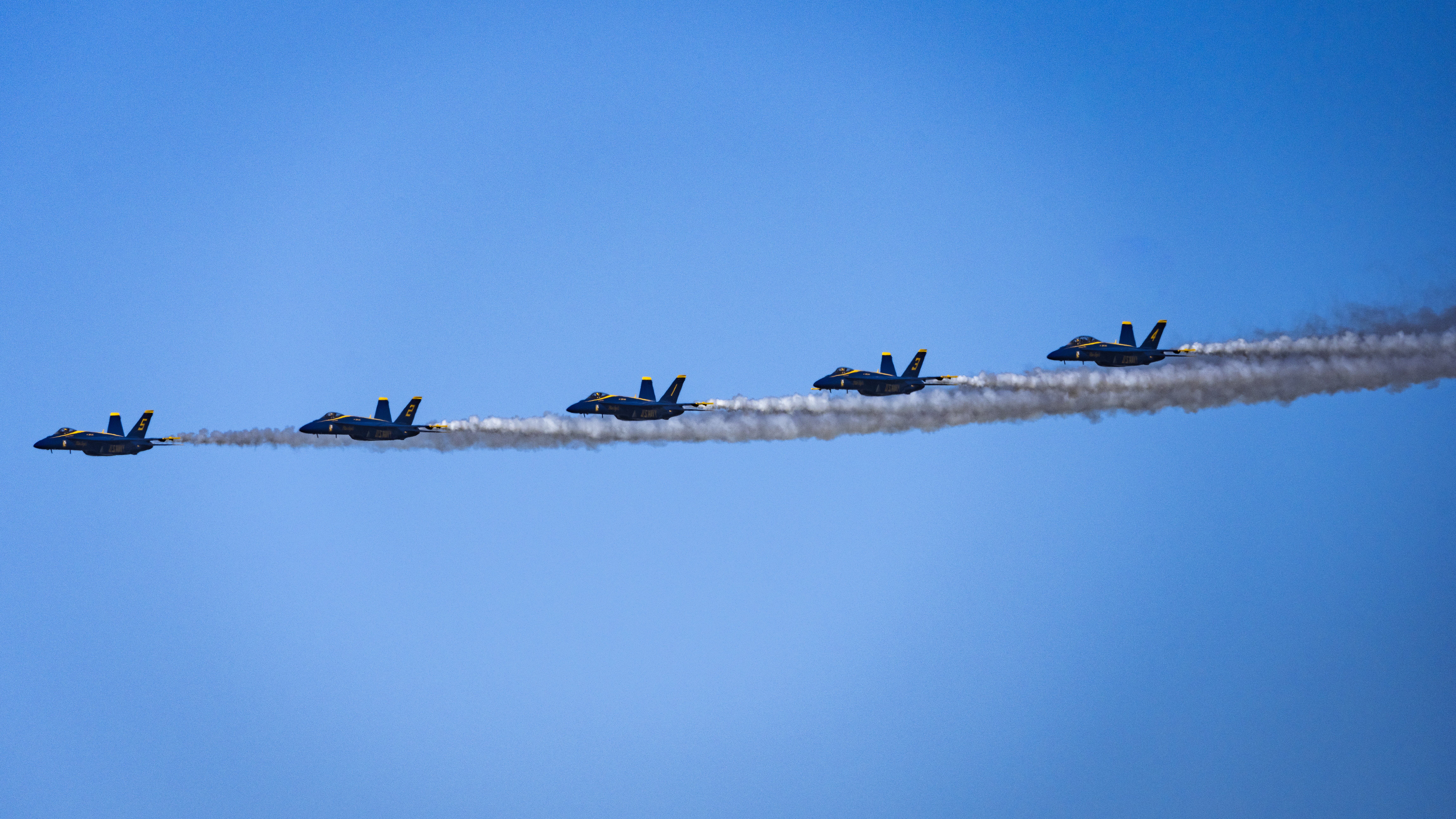 Blue Angels jets fly in a line with long white smoke trails extending behind them.