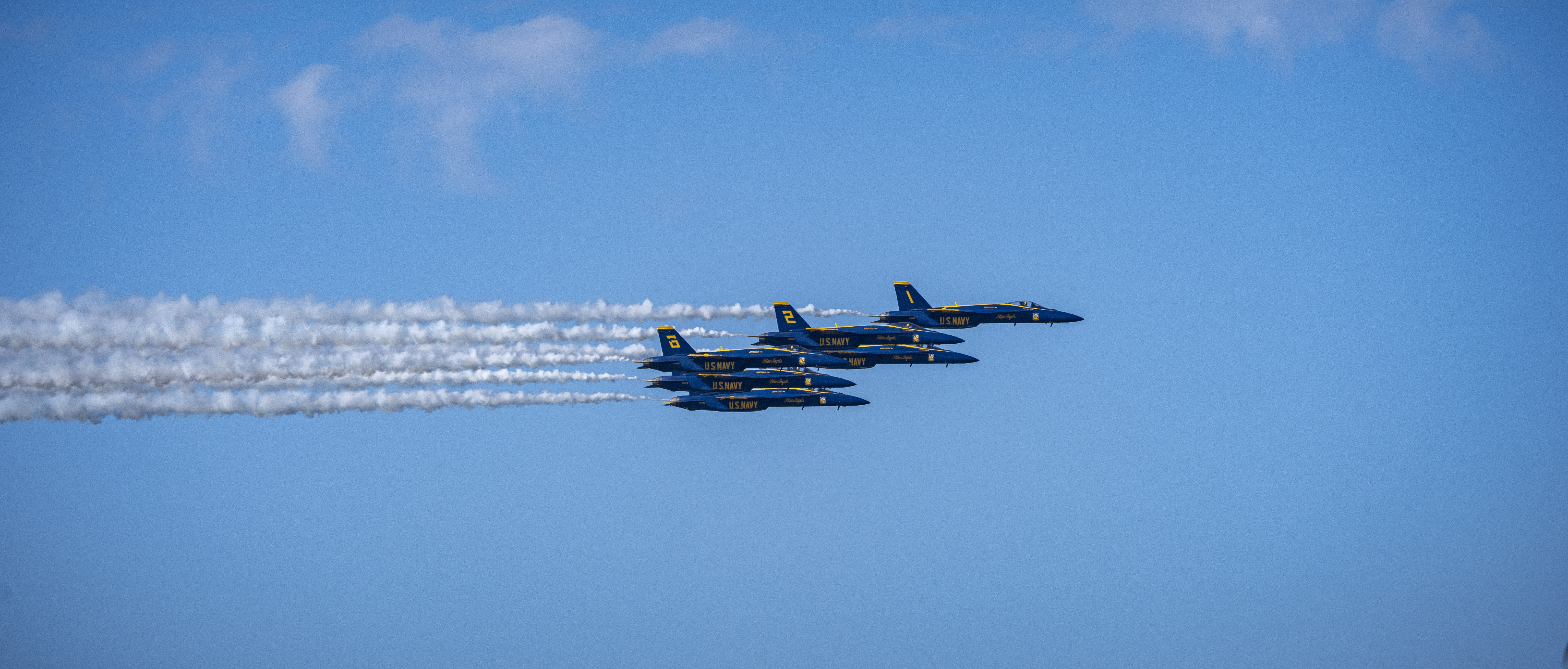 Blue Angels jets fly in close parallel formation beneath long smoke trails.