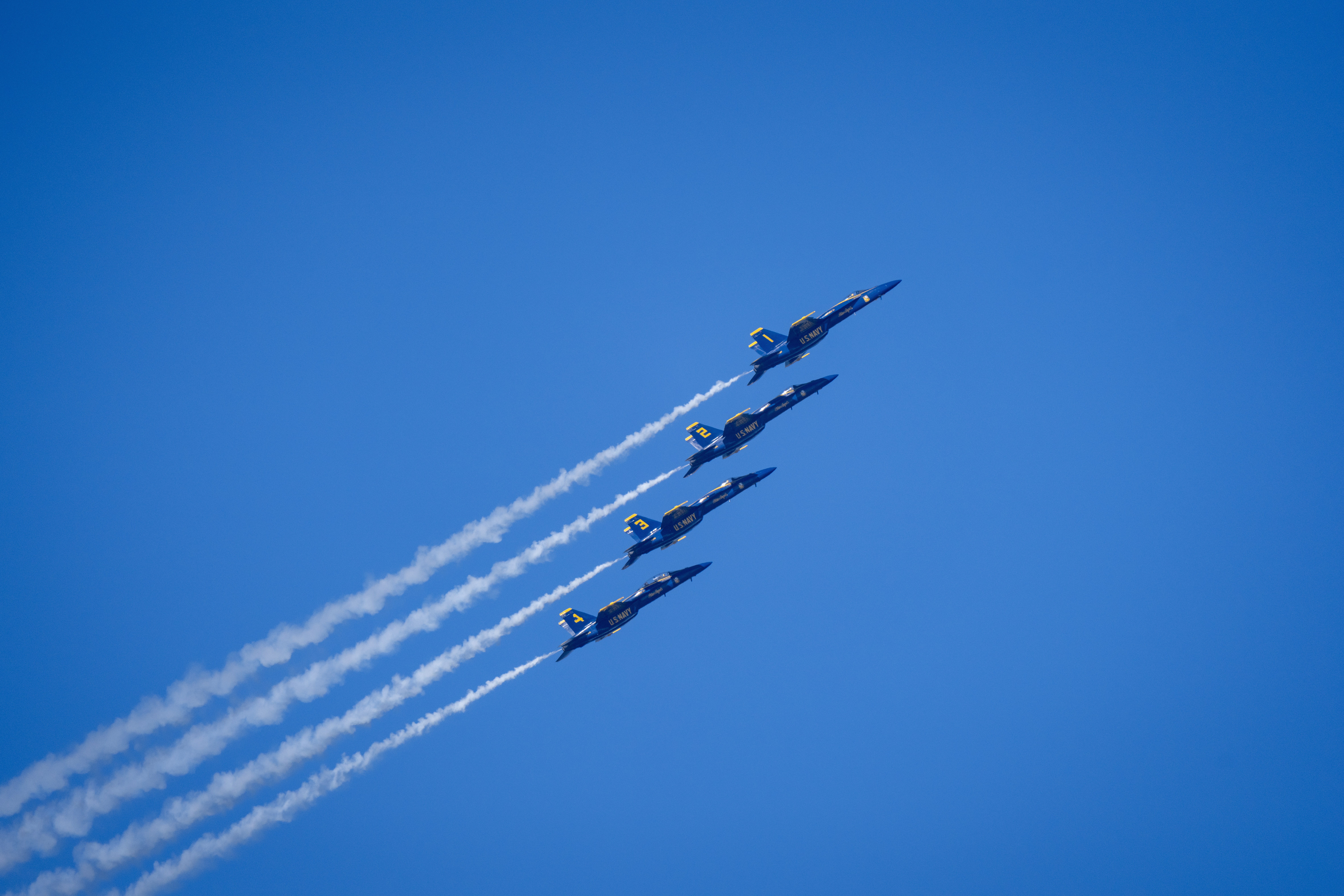 Blue Angels jets fly in a diagonal line with smoke trails across the sky.