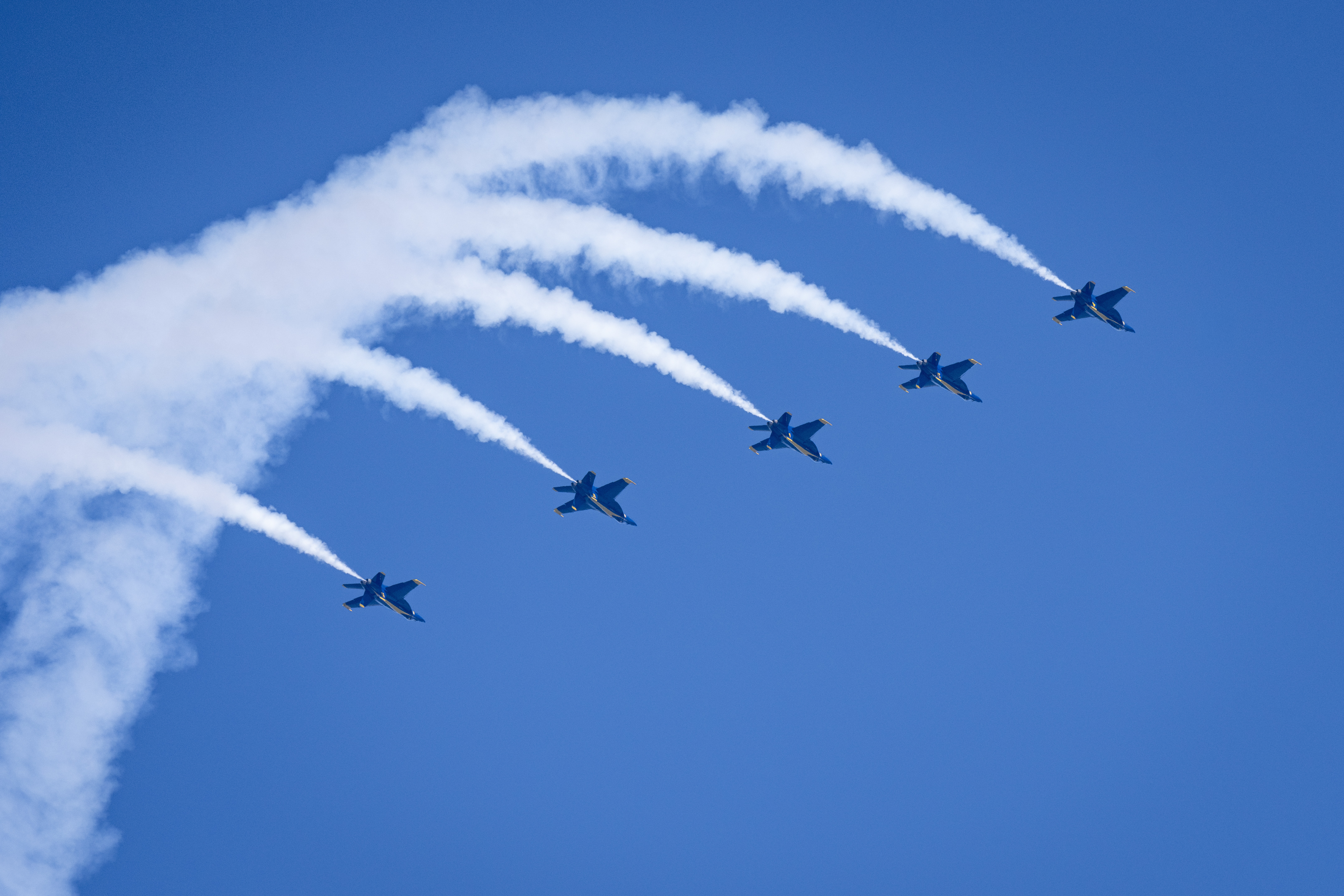 Blue Angels jets curve across the sky amid broad arcs of white smoke.