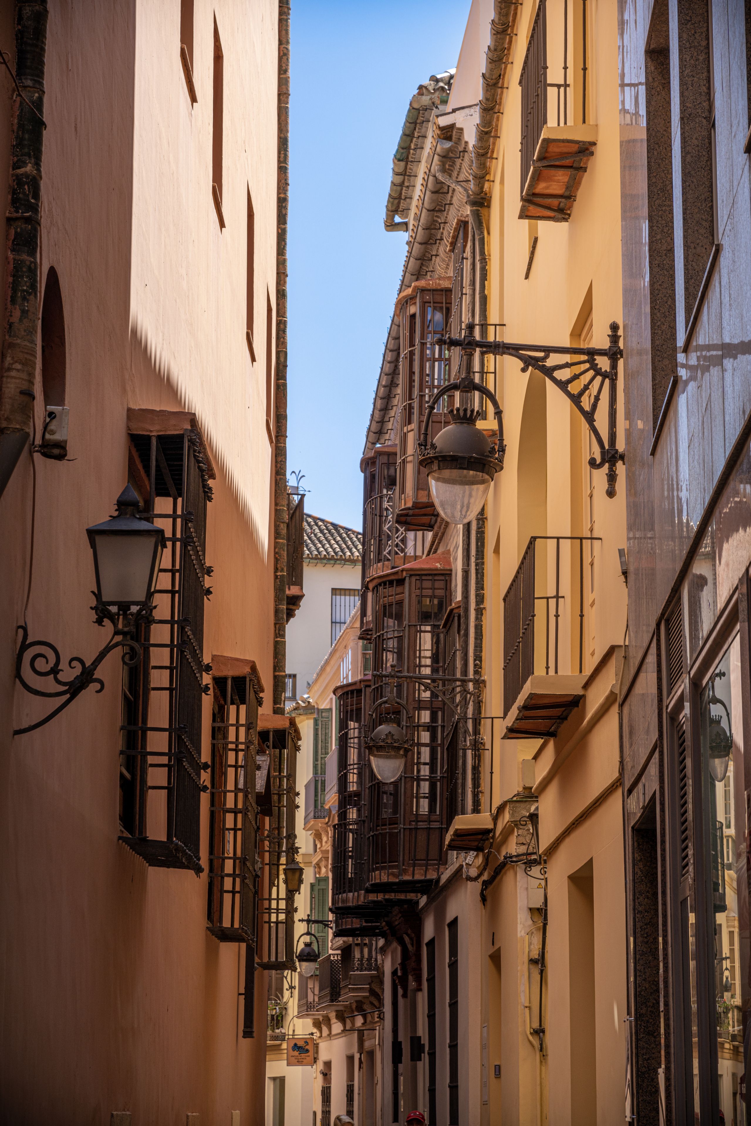 A narrow alley features buildings with decorative balconies and street lamps under a clear blue sky.