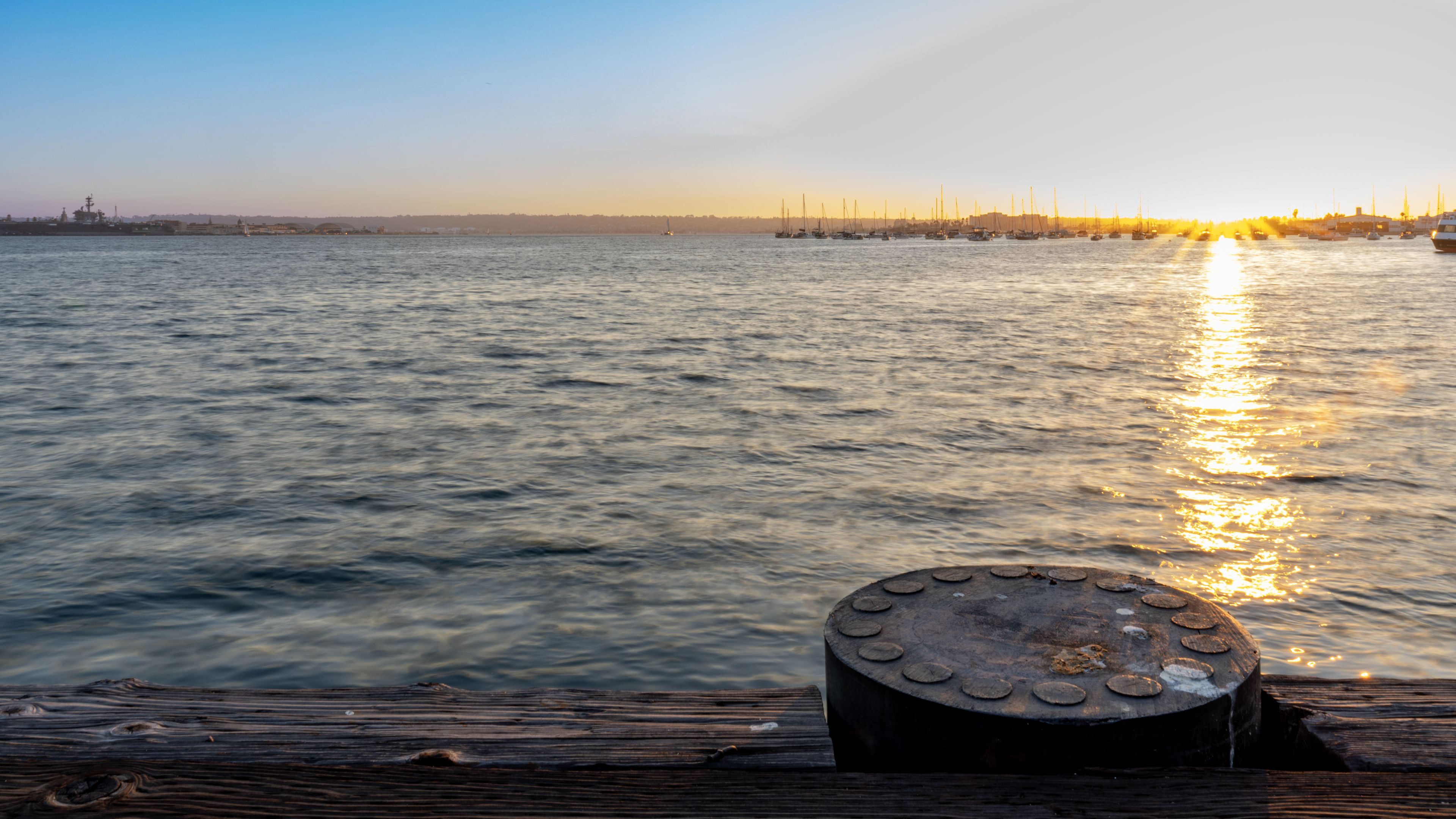 The sun sets over a calm body of water with a pier and distant sailboats.