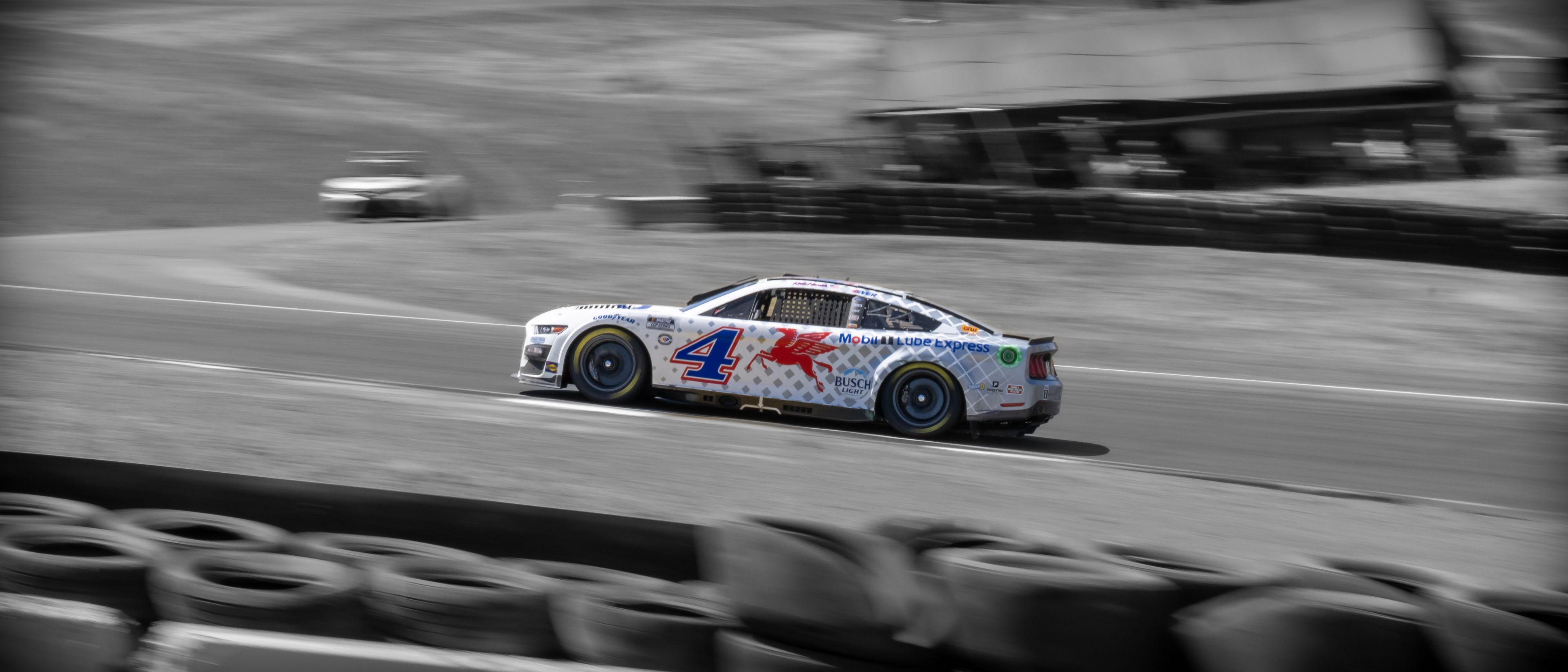 A race car speeds around a track, with tires and grass visible in the foreground.