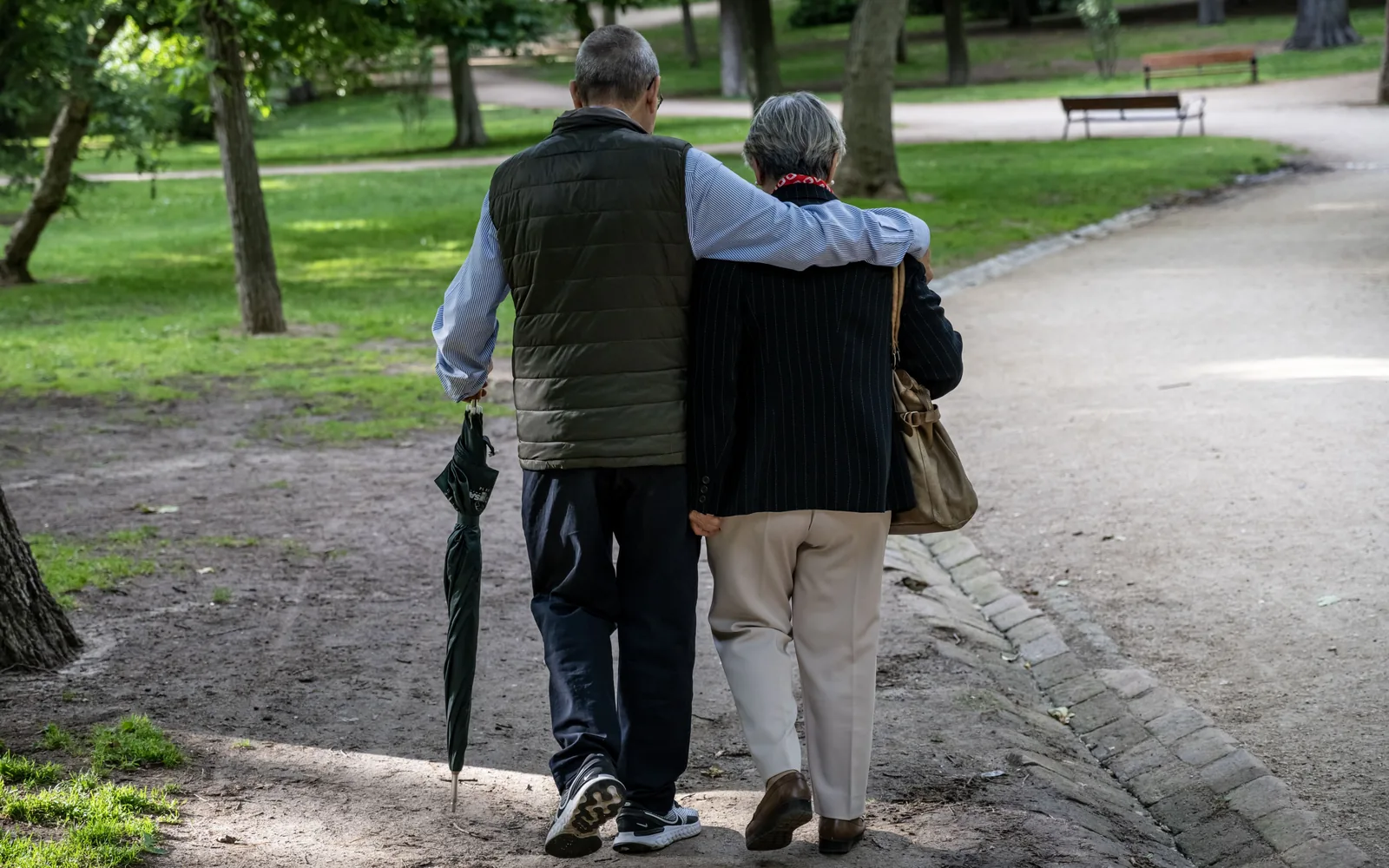 Two older adults walk away along a tree-lined park path, one with an arm around the other and holding a closed umbrella.