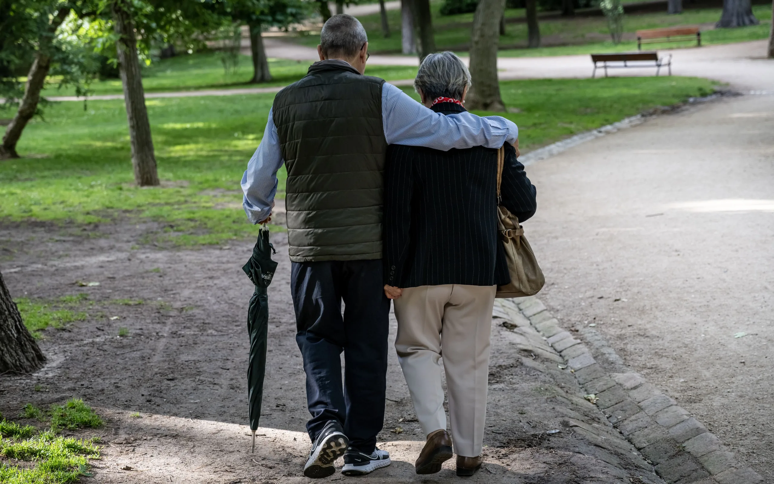 Two older adults walk away along a tree-lined park path, one with an arm around the other and holding a closed umbrella.
