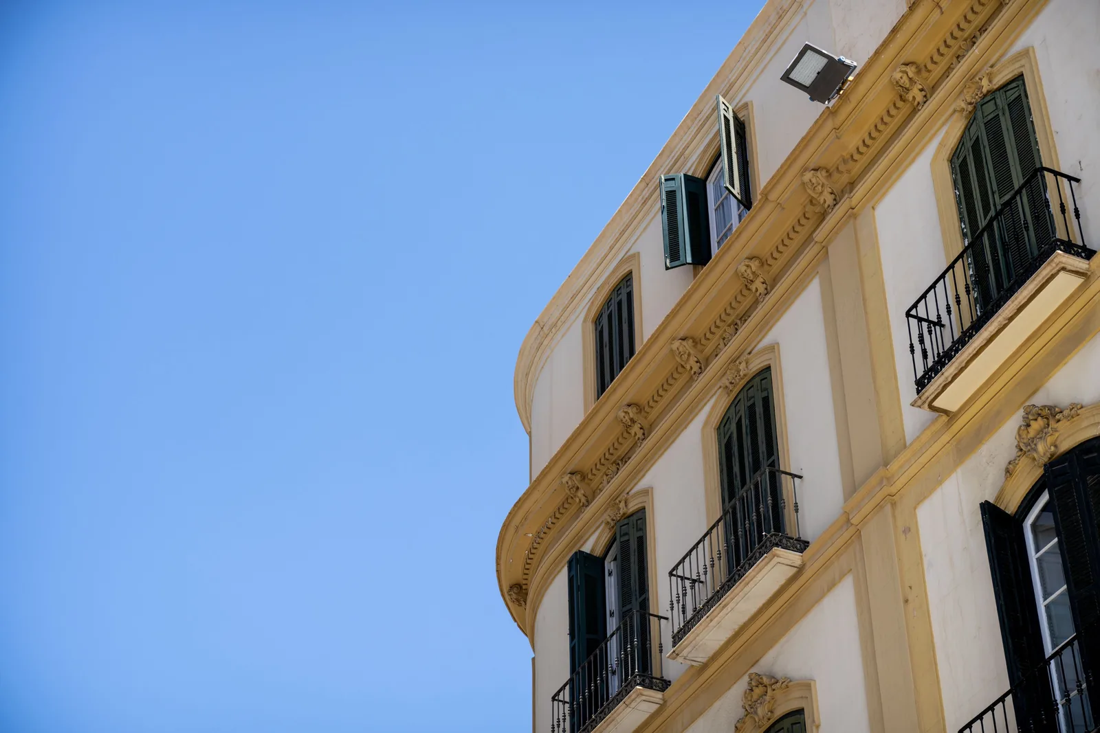 Curved cream building facade with dark open shutters, iron balconies, and decorative trim occupies the right side against blue sky.