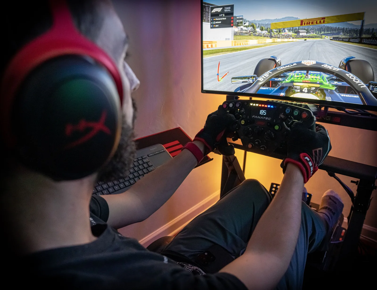 Person in a headset and racing gloves holds a simulator steering wheel facing a monitor displaying a Formula 1 racing game.