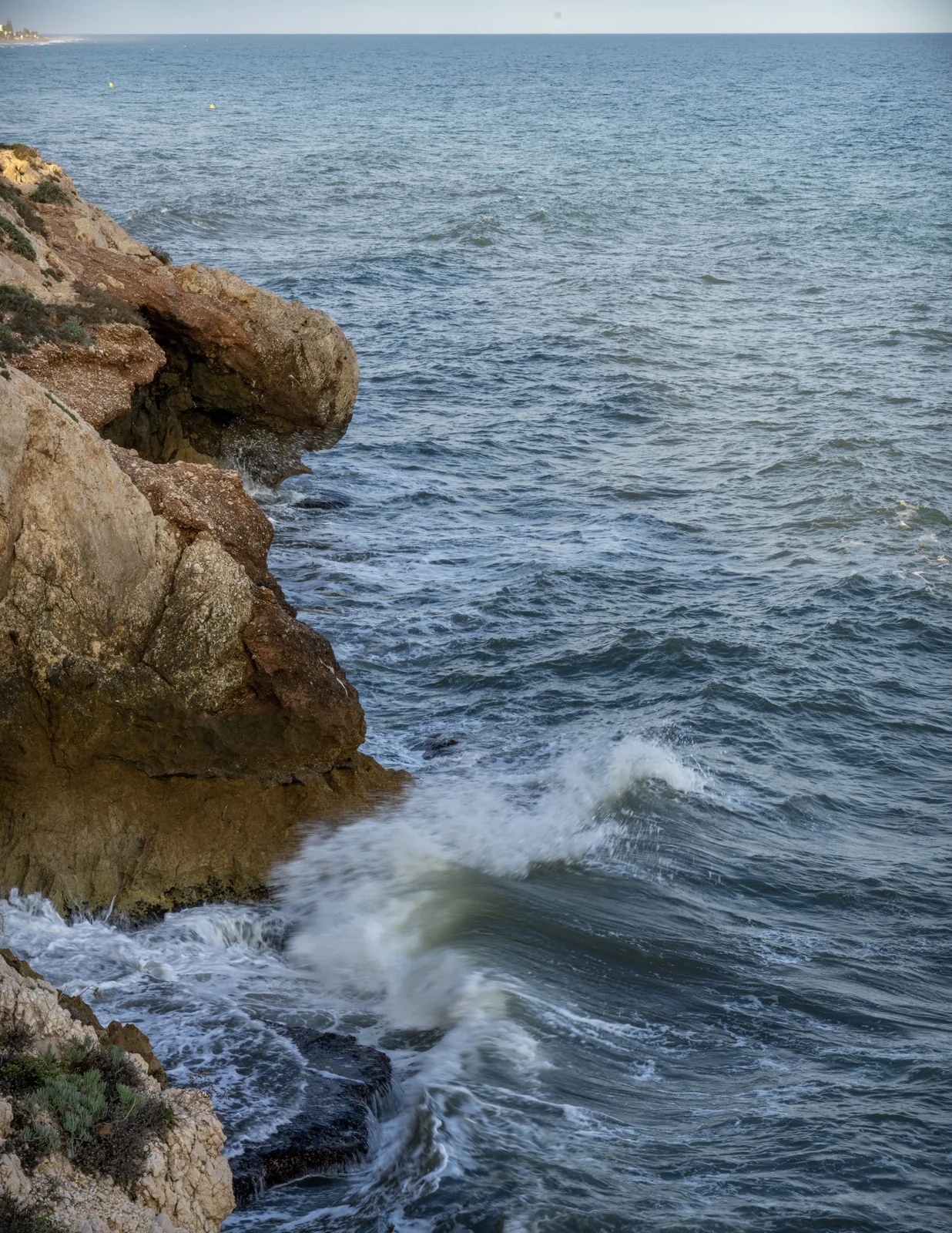 Rough coastal rocks occupy the left side as white water moves below, with open sea, yellow buoys, and a distant shoreline beyond.