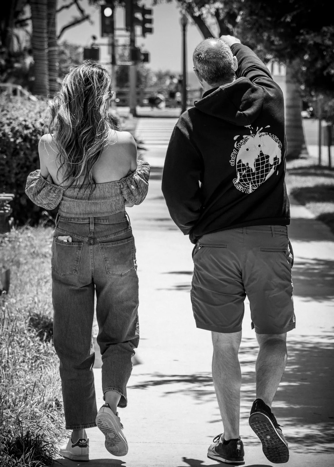 Two people walk side by side along a tree-lined sidewalk, seen from behind in a black-and-white photograph.