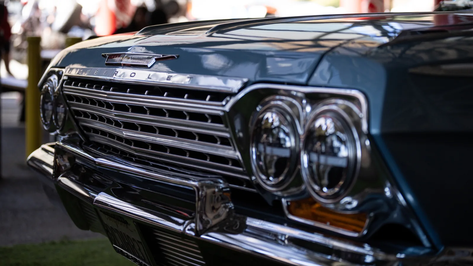 Close-up of a blue Chevrolet Impala front end with twin round headlights, chrome grille, hood emblem, and blurred people behind it.