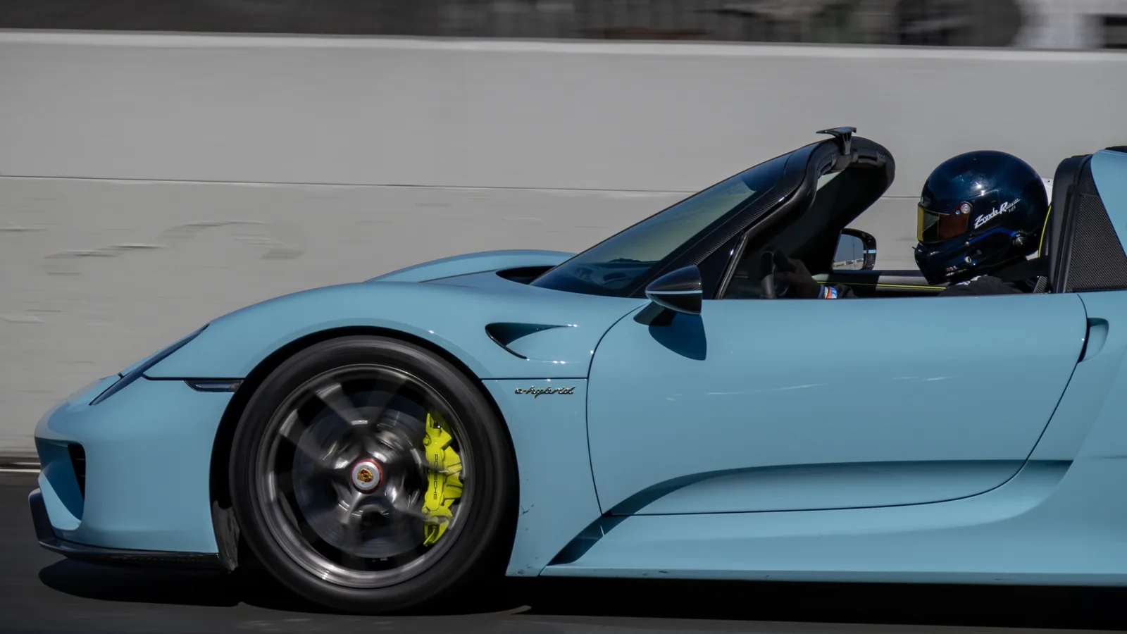 Side view of a light blue open-top Porsche sports car with a helmeted driver, blurred front wheel, and bright yellow brake caliper.