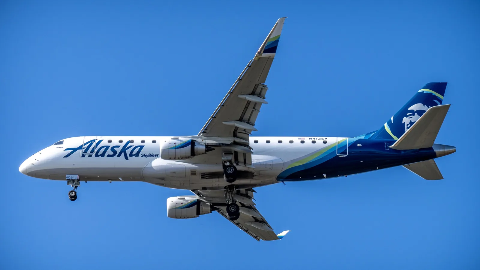 Alaska Airlines aircraft seen from below with landing gear extended, swept wings, and blue-and-green tail markings against blue sky.