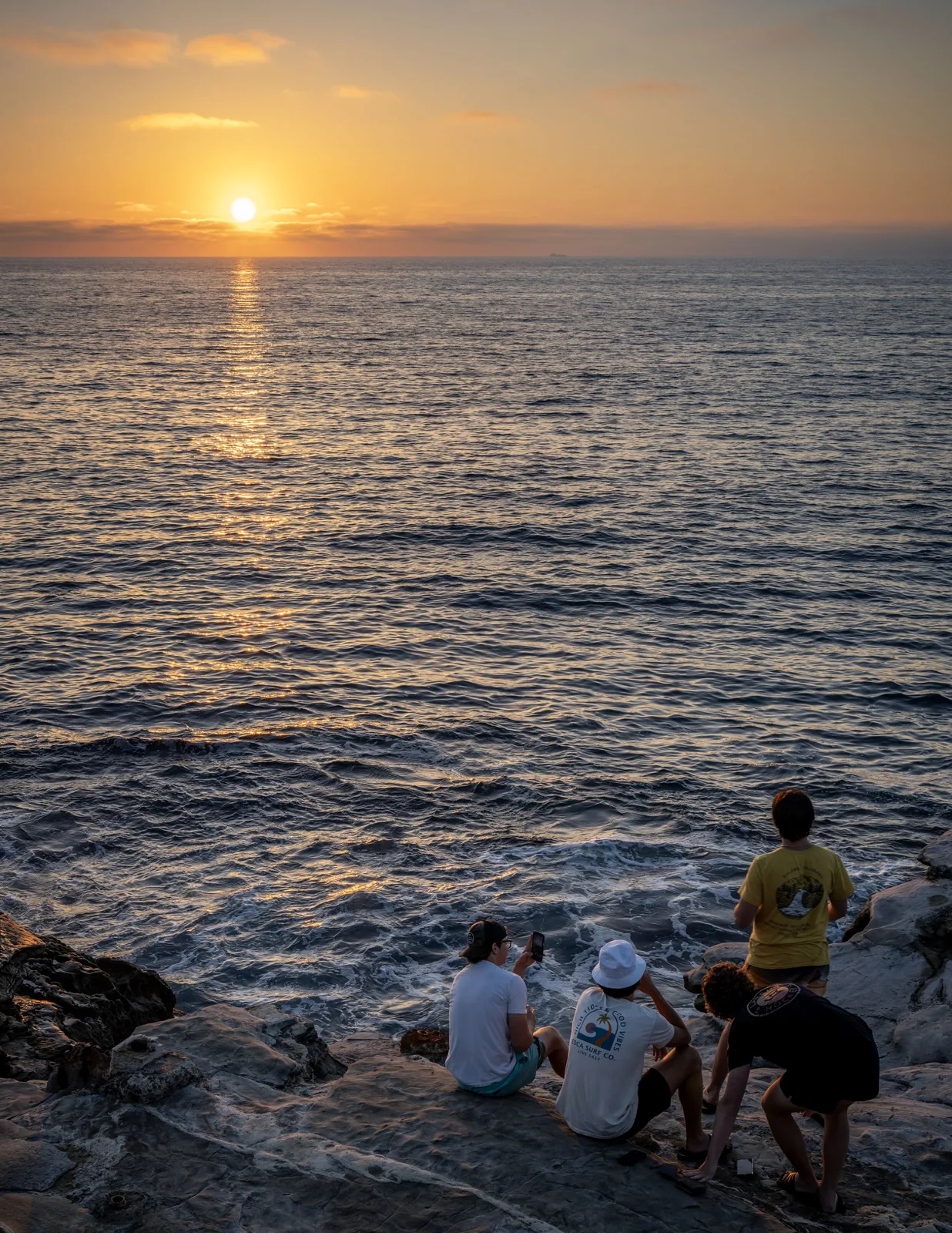 Several people sit and stand on coastal rocks beside choppy water, facing the low sun over the ocean.