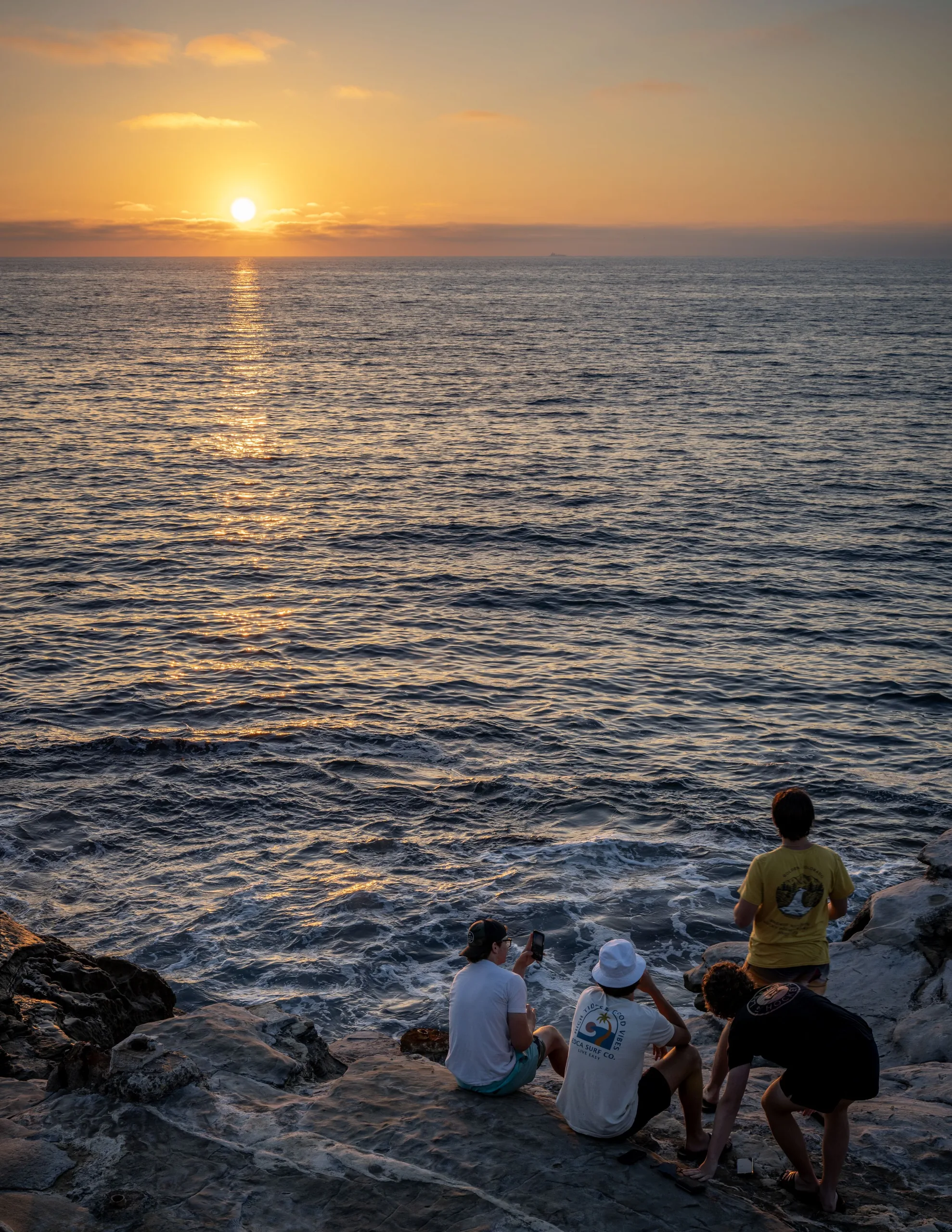 Several people sit and stand on coastal rocks beside choppy water, facing the low sun over the ocean.
