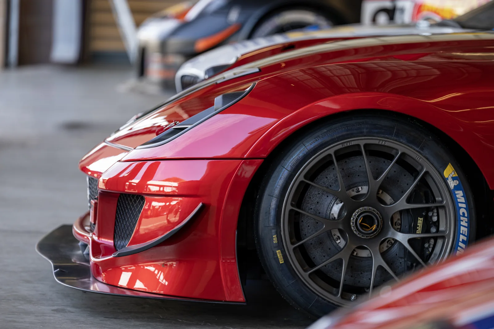 Close-up of the front quarter of a red sports car, showing aerodynamic bodywork, a dark multi-spoke wheel, brake rotor, and Michelin tire.