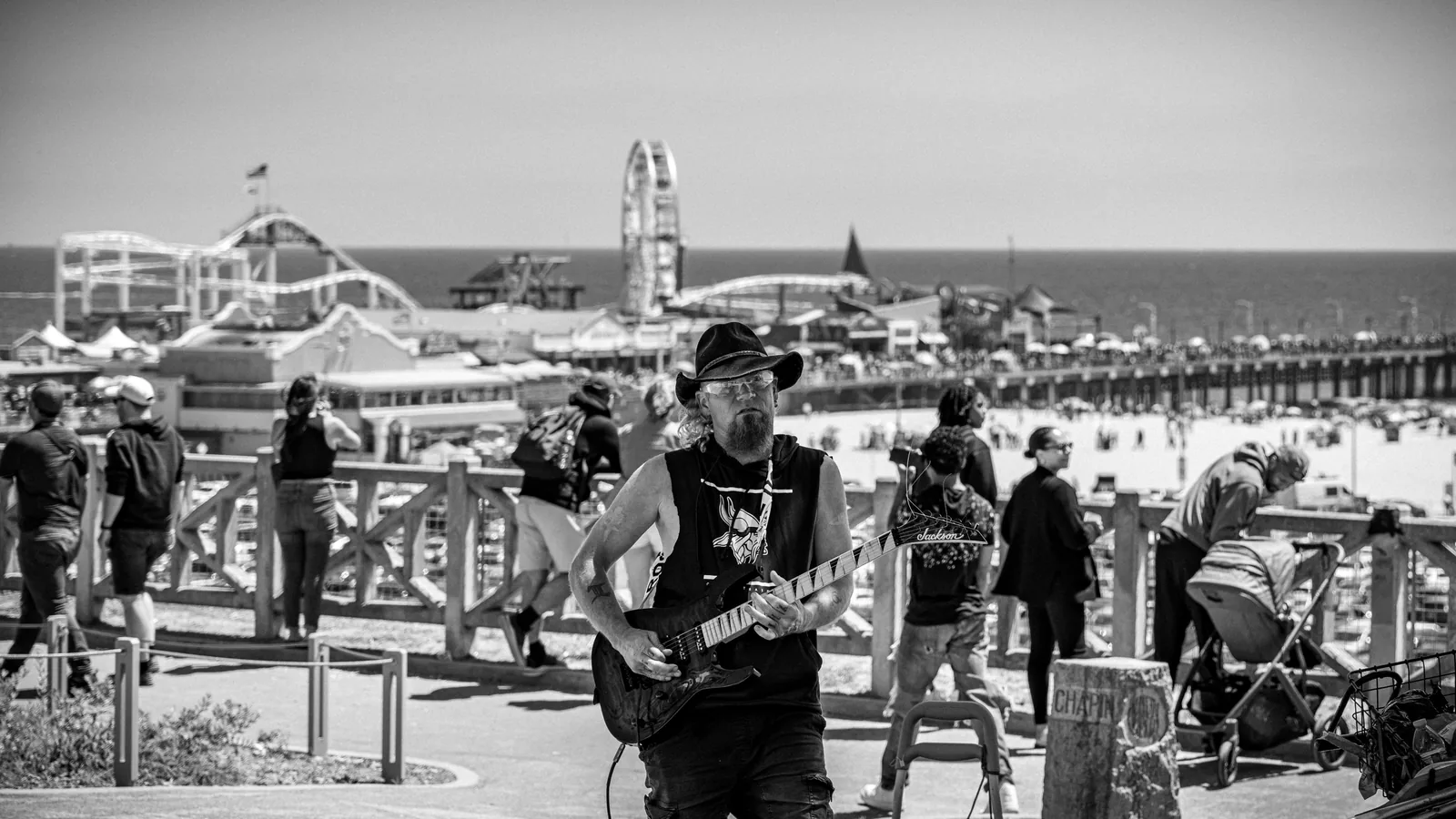 Man in a cowboy hat plays an electric guitar on a raised walkway, with a beach, amusement rides, and pier behind him.