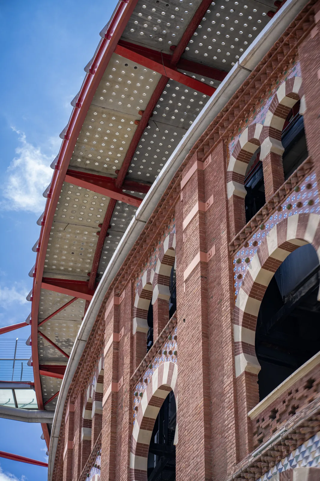 Upward view of a red-brick building with striped arches and patterned tilework beneath a perforated metal canopy.