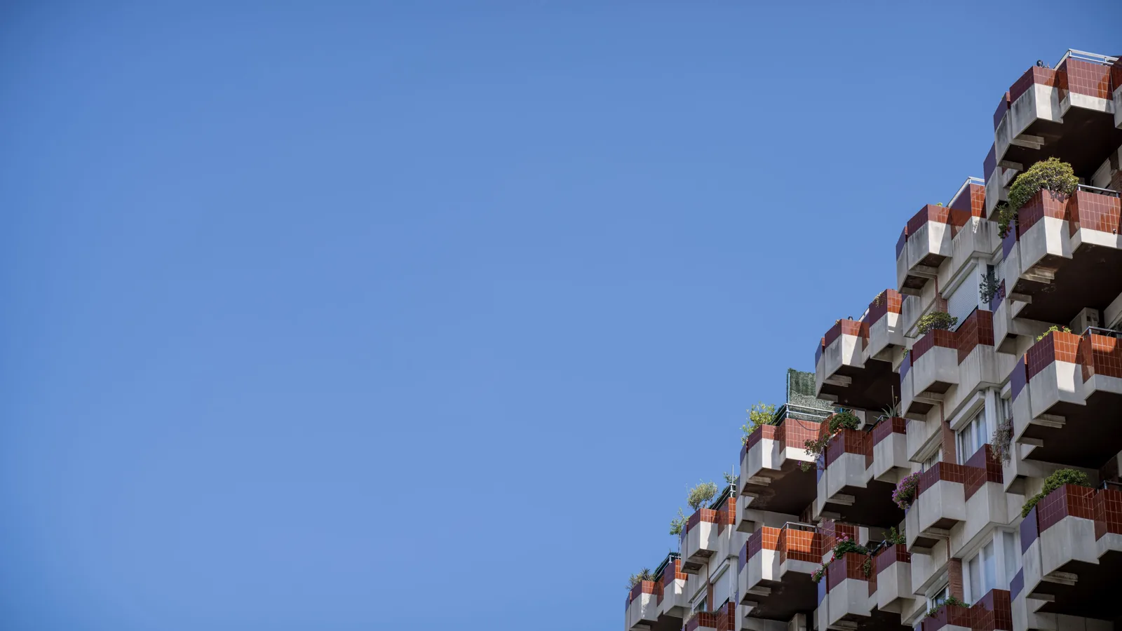 Modern apartment building with projecting balconies and red tile sections occupying the right side against a broad blue sky.