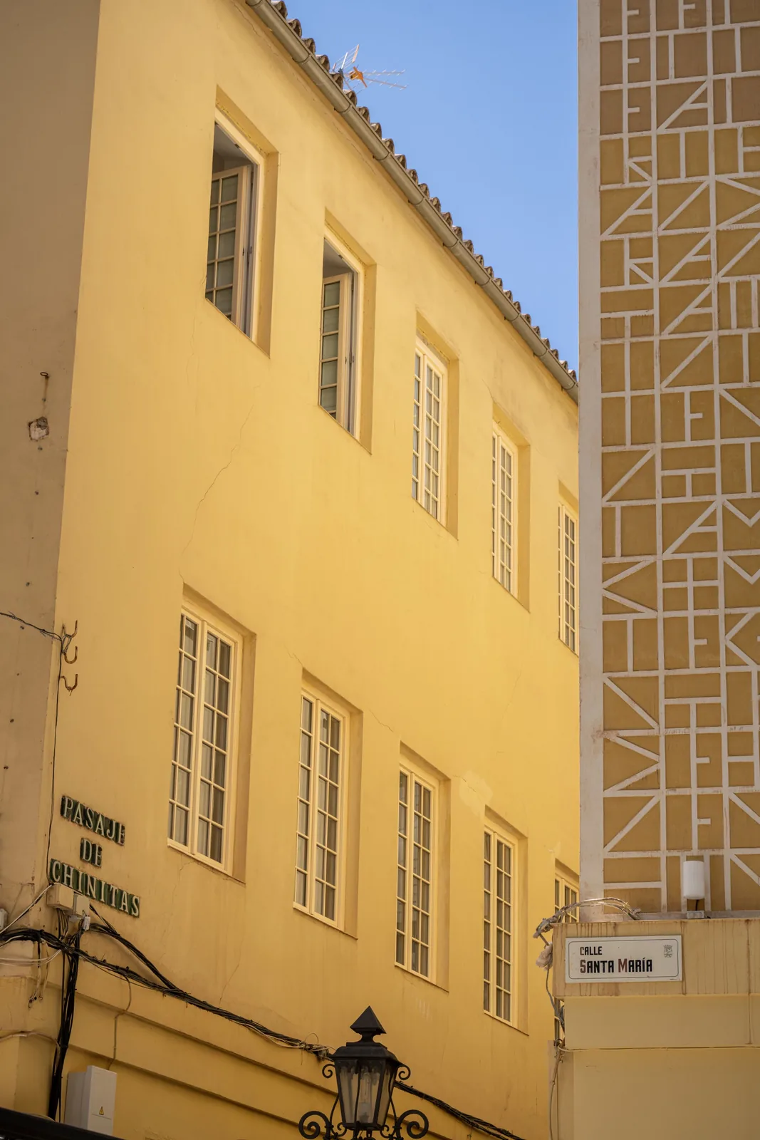 Yellow building with tall multi-pane windows stands beside a geometric patterned wall, with street signs for Pasaje de Chinitas and Calle Santa María.