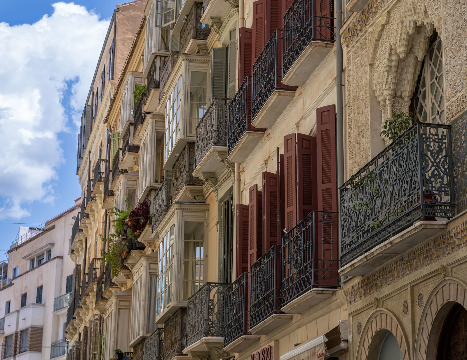 Ornate building facades rise along a narrow street, lined with wrought-iron balconies, projecting windows, shutters, and carved stonework beneath blue sky.