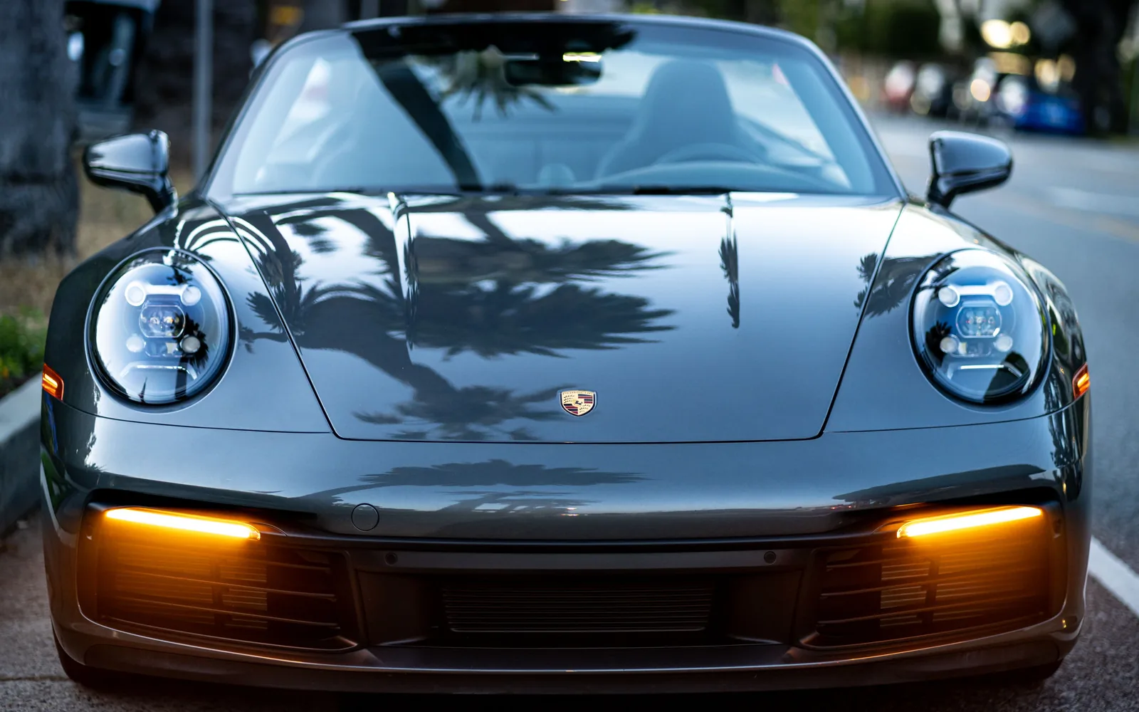 Front view of a Porsche sports car with round headlights, amber lower lights, and palm tree reflections across its hood.