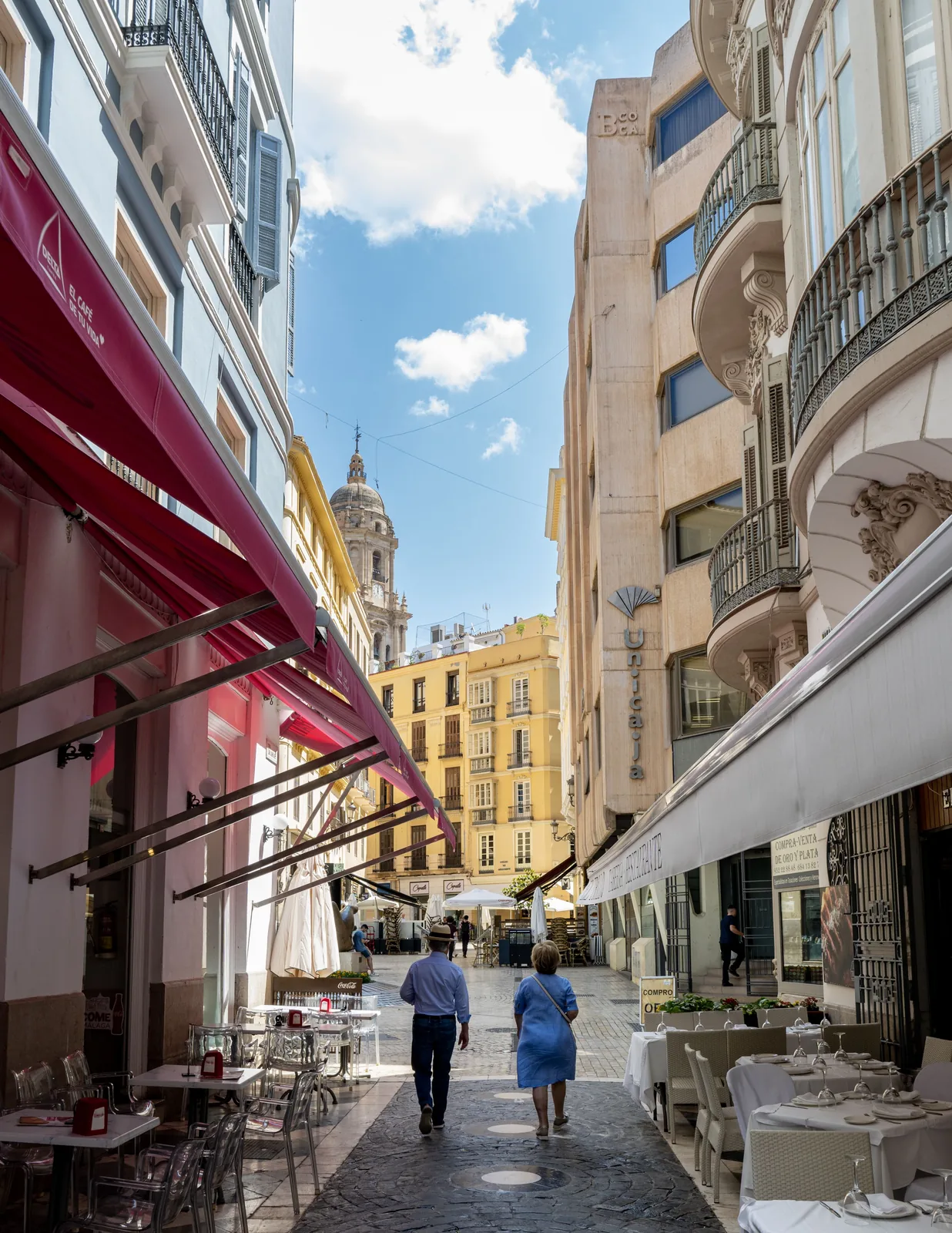 Two pedestrians walk through a narrow paved street lined with restaurants and balconies, with a domed tower visible ahead.