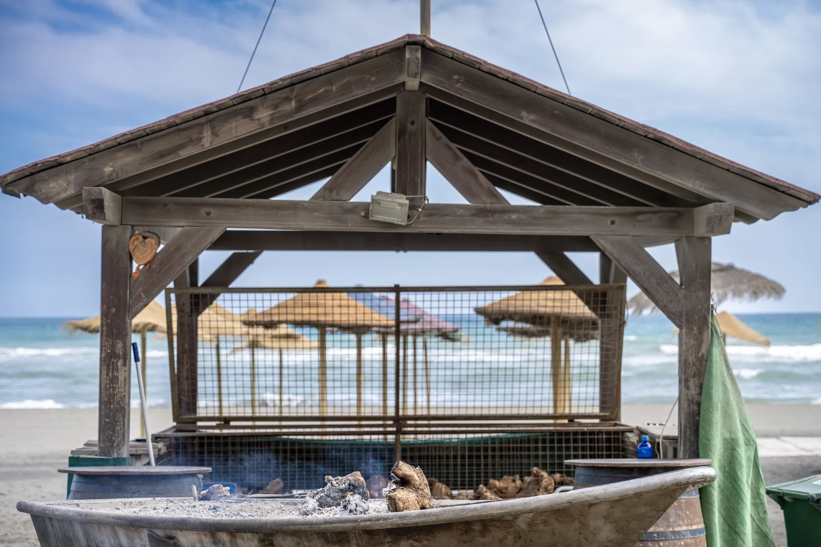 A wooden roofed beach shelter frames straw umbrellas and the sea, while a large metal brazier with ash and charred wood fills the foreground.