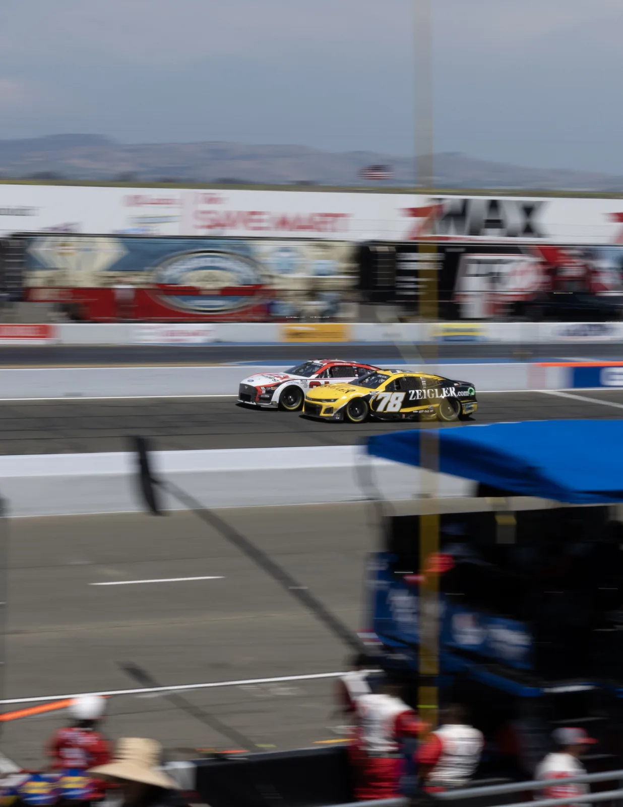 Two stock cars travel side by side on a racetrack, with blurred pit equipment and mountains in the background.