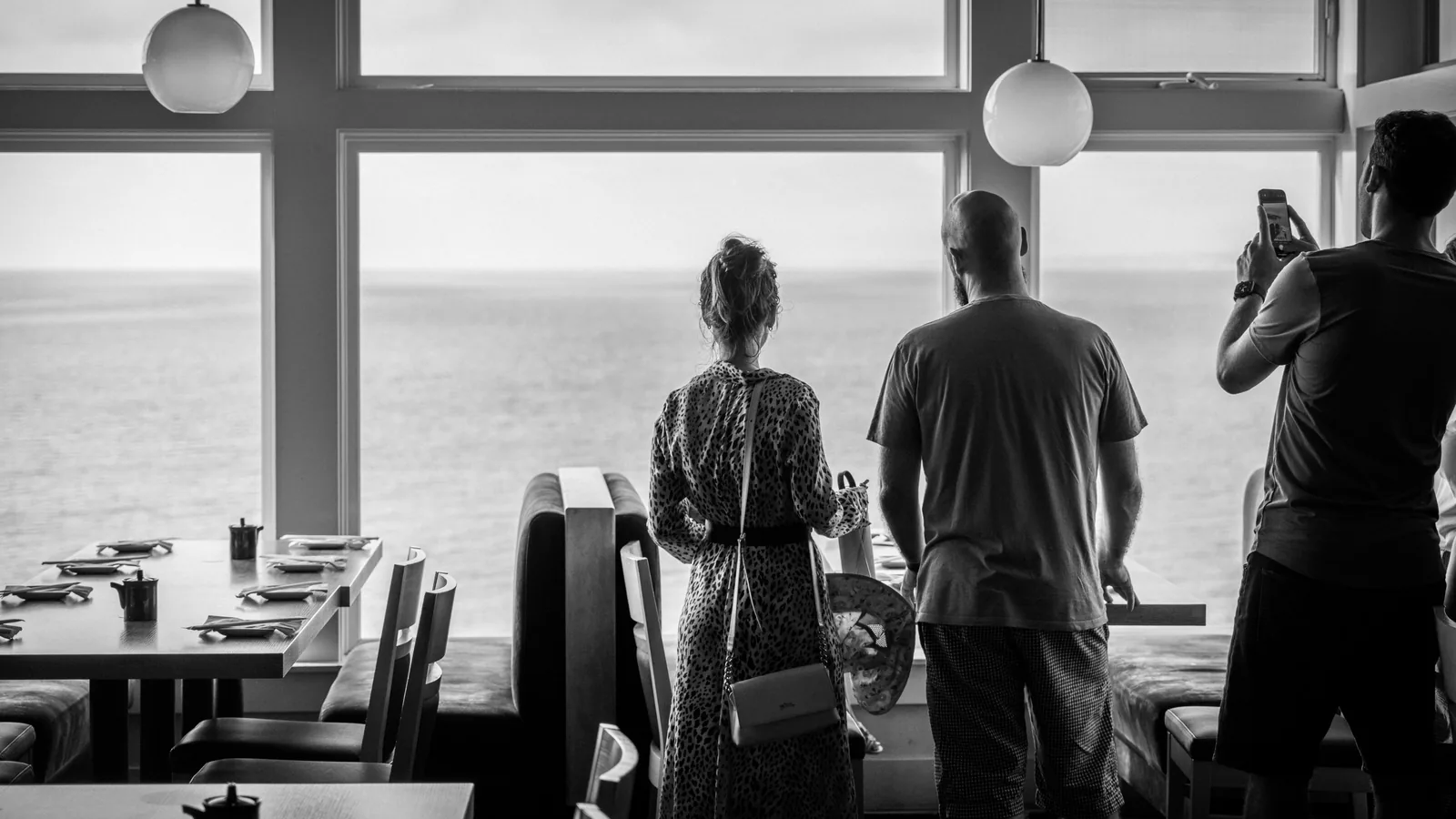 Three people stand inside a restaurant facing the ocean through large windows, while set dining tables fill the foreground.