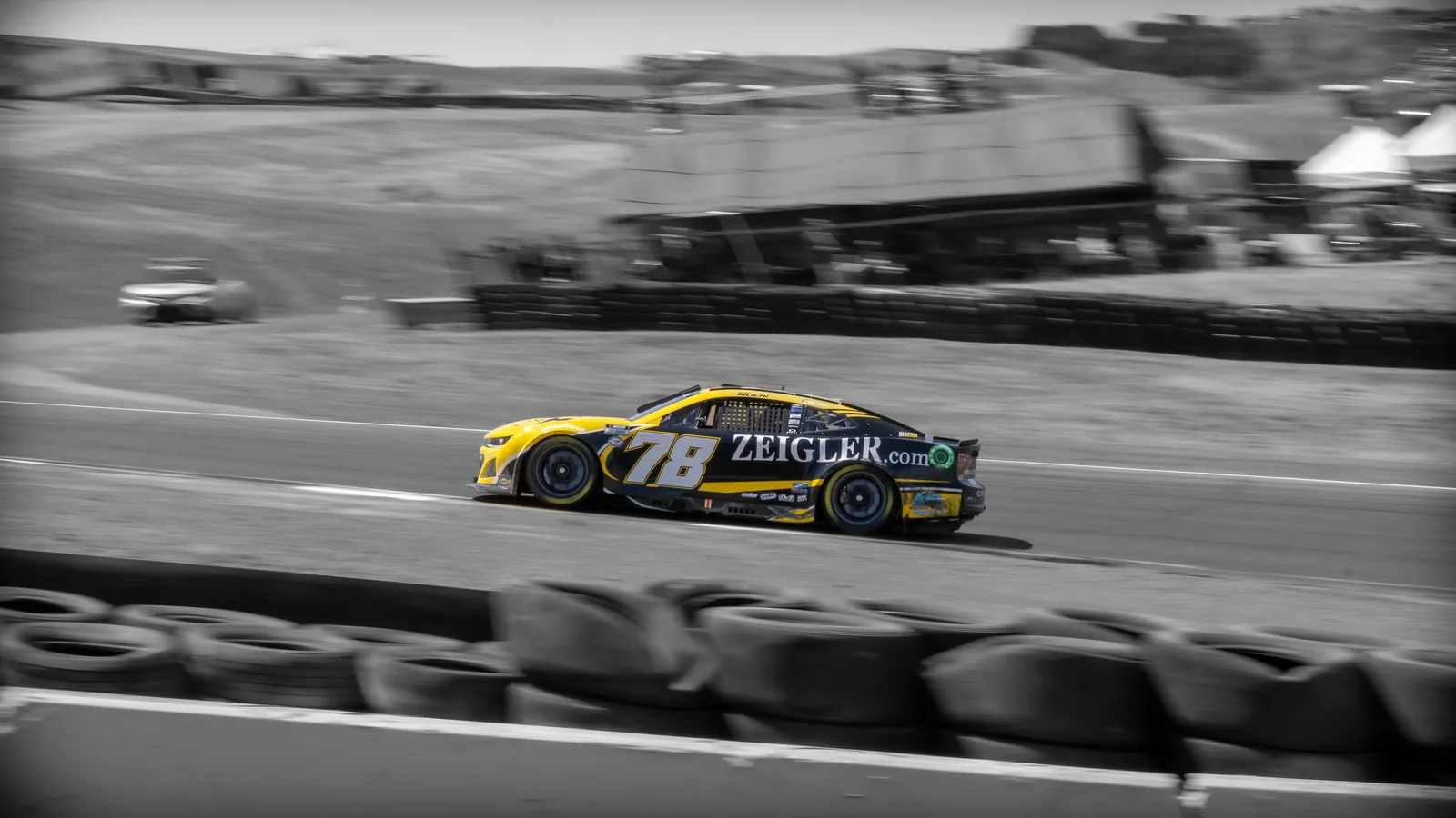 Yellow-and-black stock car numbered 78 drives on a racetrack, isolated in color against motion-blurred black-and-white surroundings and tire barriers.