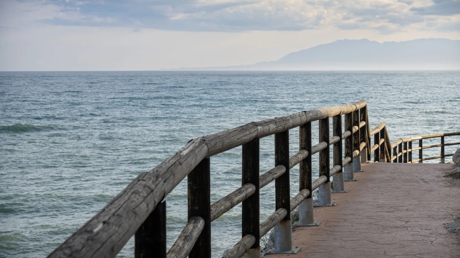 A paved coastal walkway curves along the water behind rustic wooden railings, with open sea and a hazy mountainous shoreline beyond.