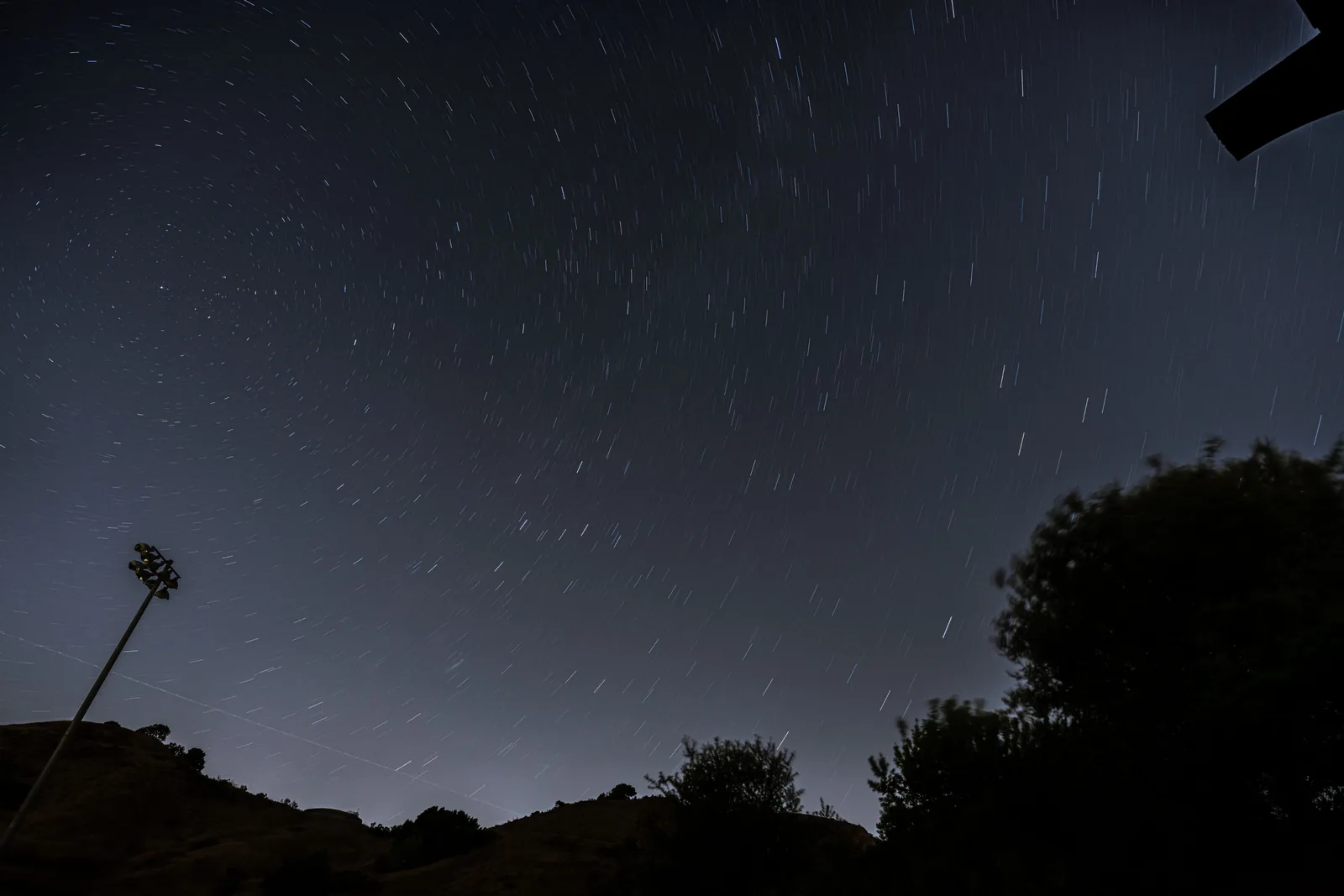 Long-exposure night sky with curved star trails above silhouetted hills and trees, with a tall light pole at the left edge.