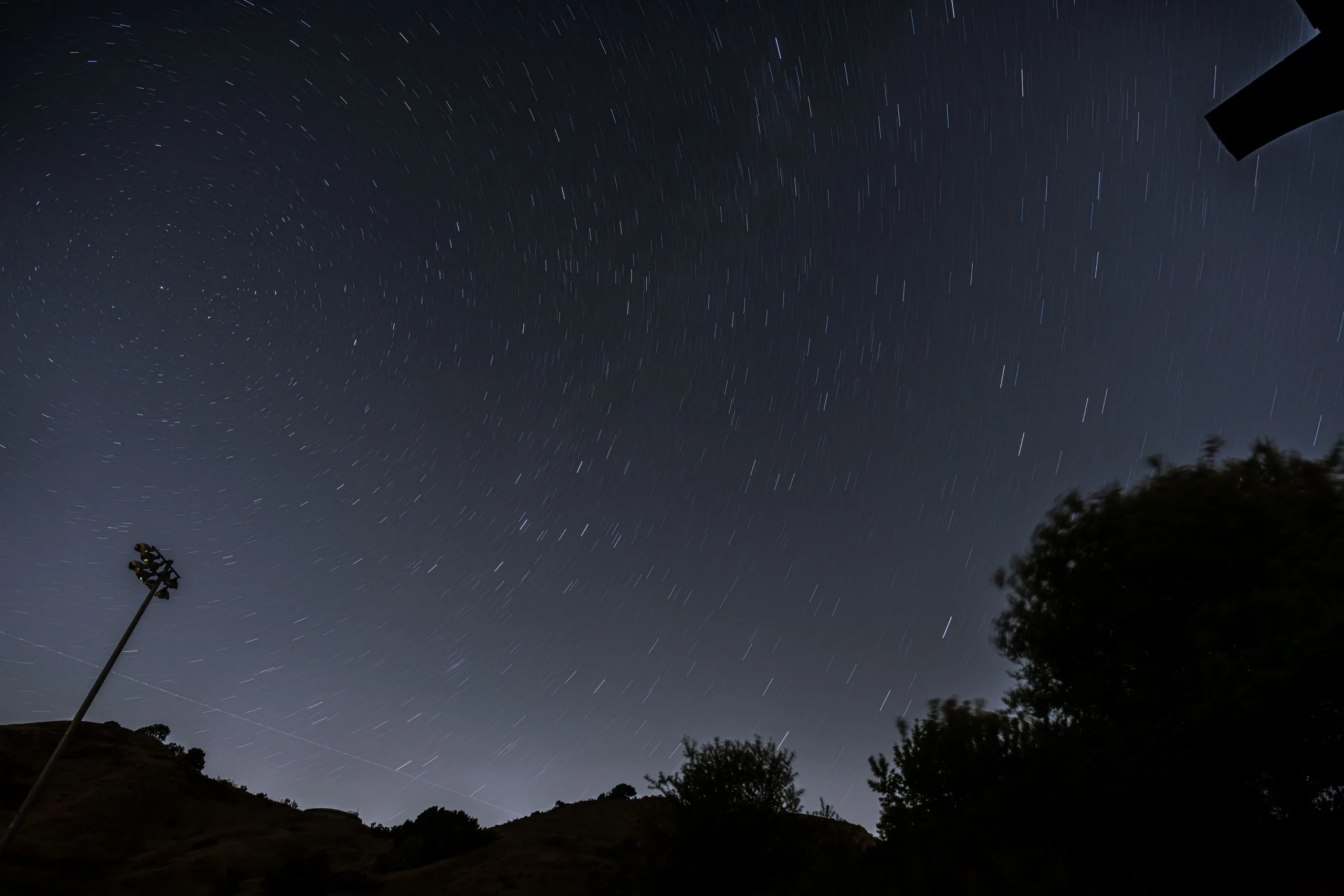Long-exposure night sky with curved star trails above silhouetted hills and trees, with a tall light pole at the left edge.