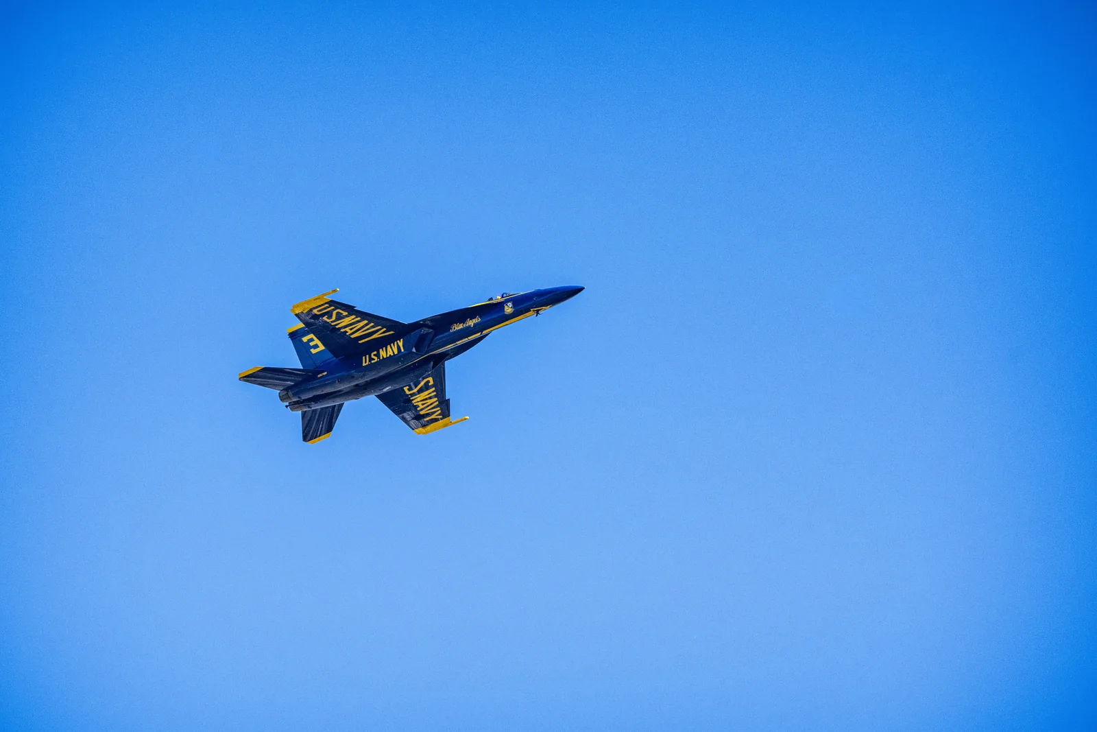 A Blue Angels jet banks across the sky with a faint smoke trail behind it.