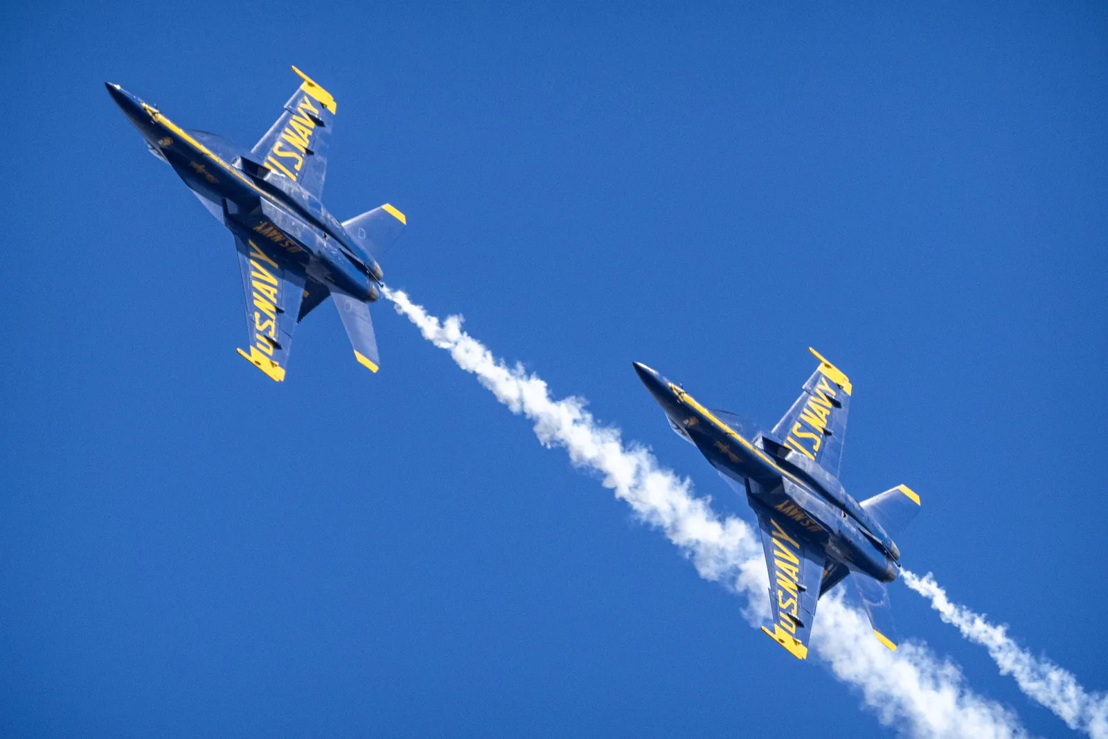 Two Blue Angels jets fly close together with smoke trailing behind them.