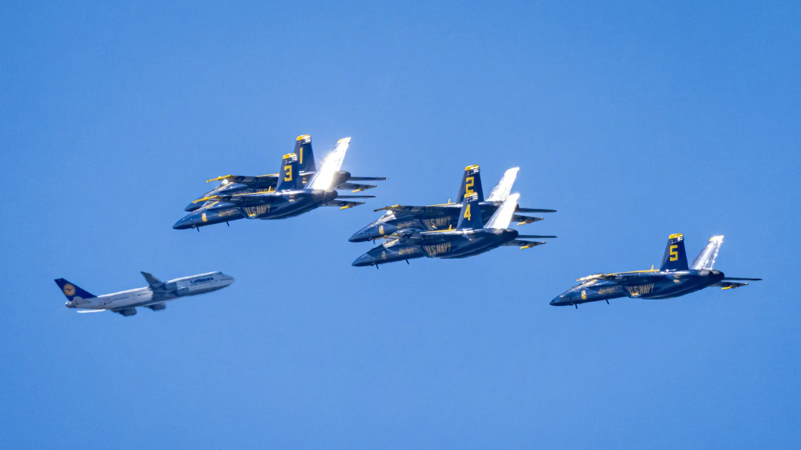 Five Blue Angels jets fly in formation beside a gray aircraft.