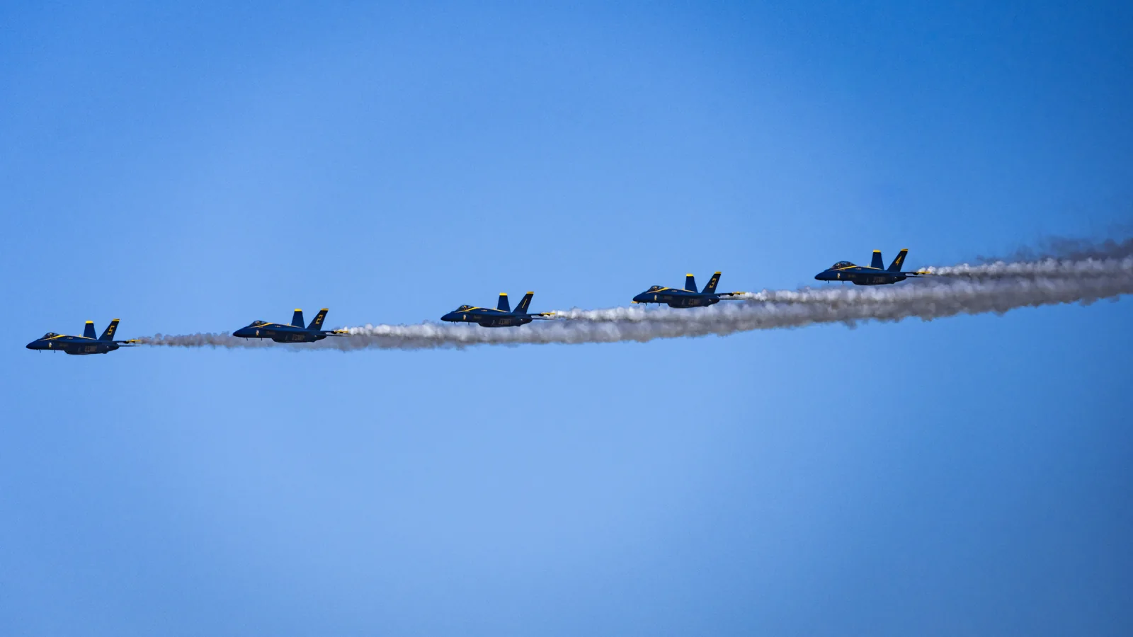 Blue Angels jets fly in a line with long white smoke trails extending behind them.
