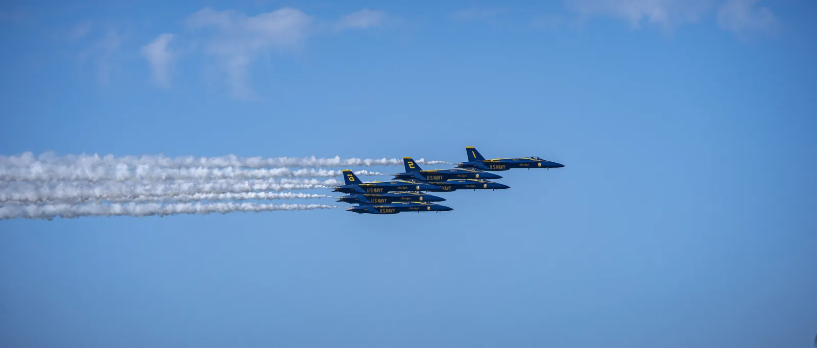Blue Angels jets fly in close parallel formation beneath long smoke trails.