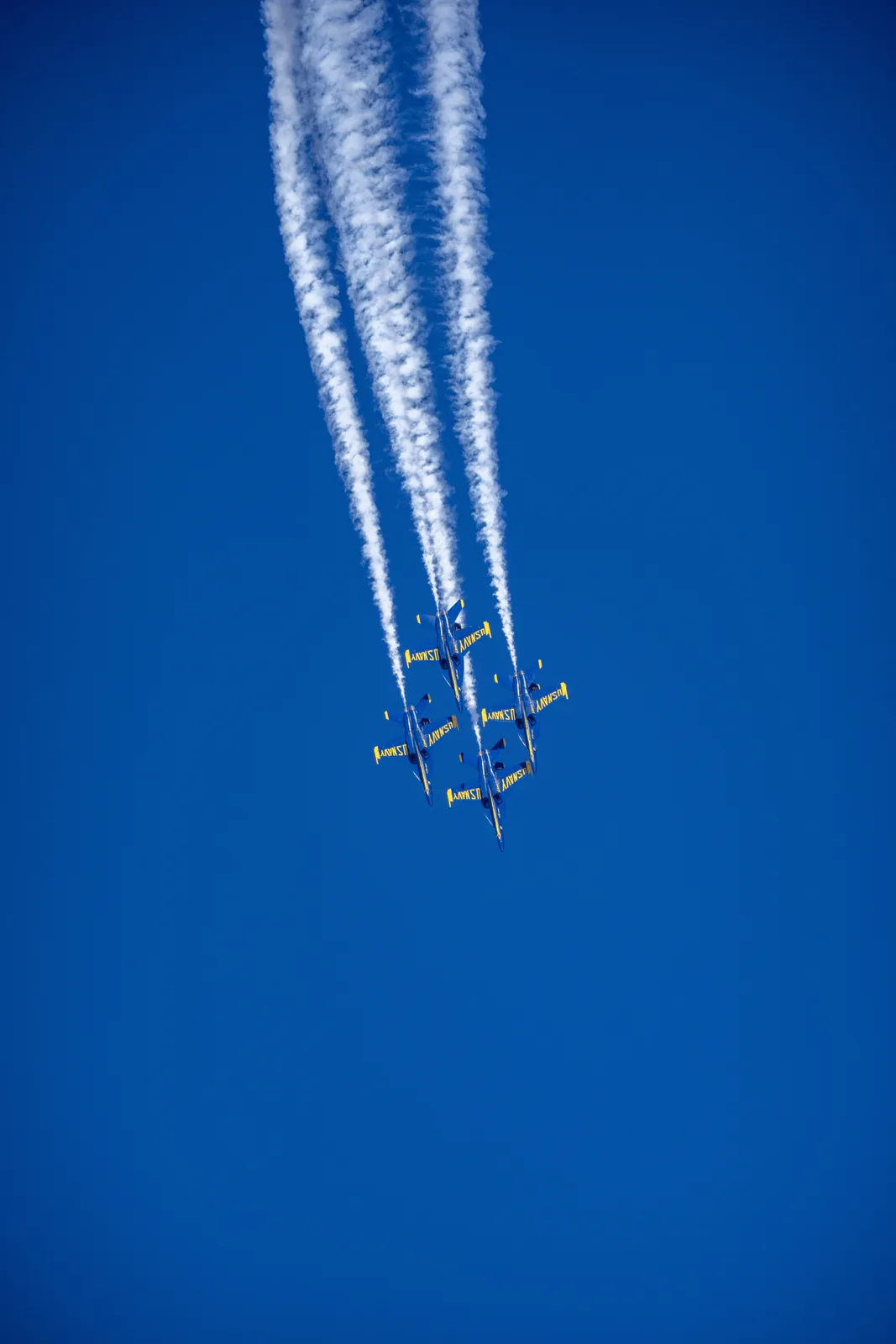 Blue Angels jets pass beneath several long parallel smoke trails.