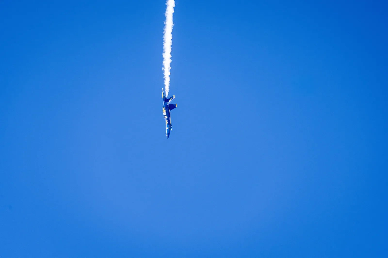 A distant Blue Angels jet passes beside a column of smoke.