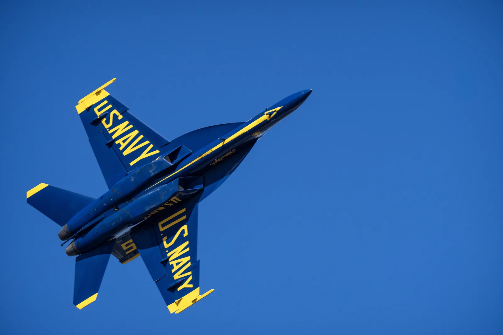A Blue Angels jet passes overhead with U.S. Navy markings visible underneath.